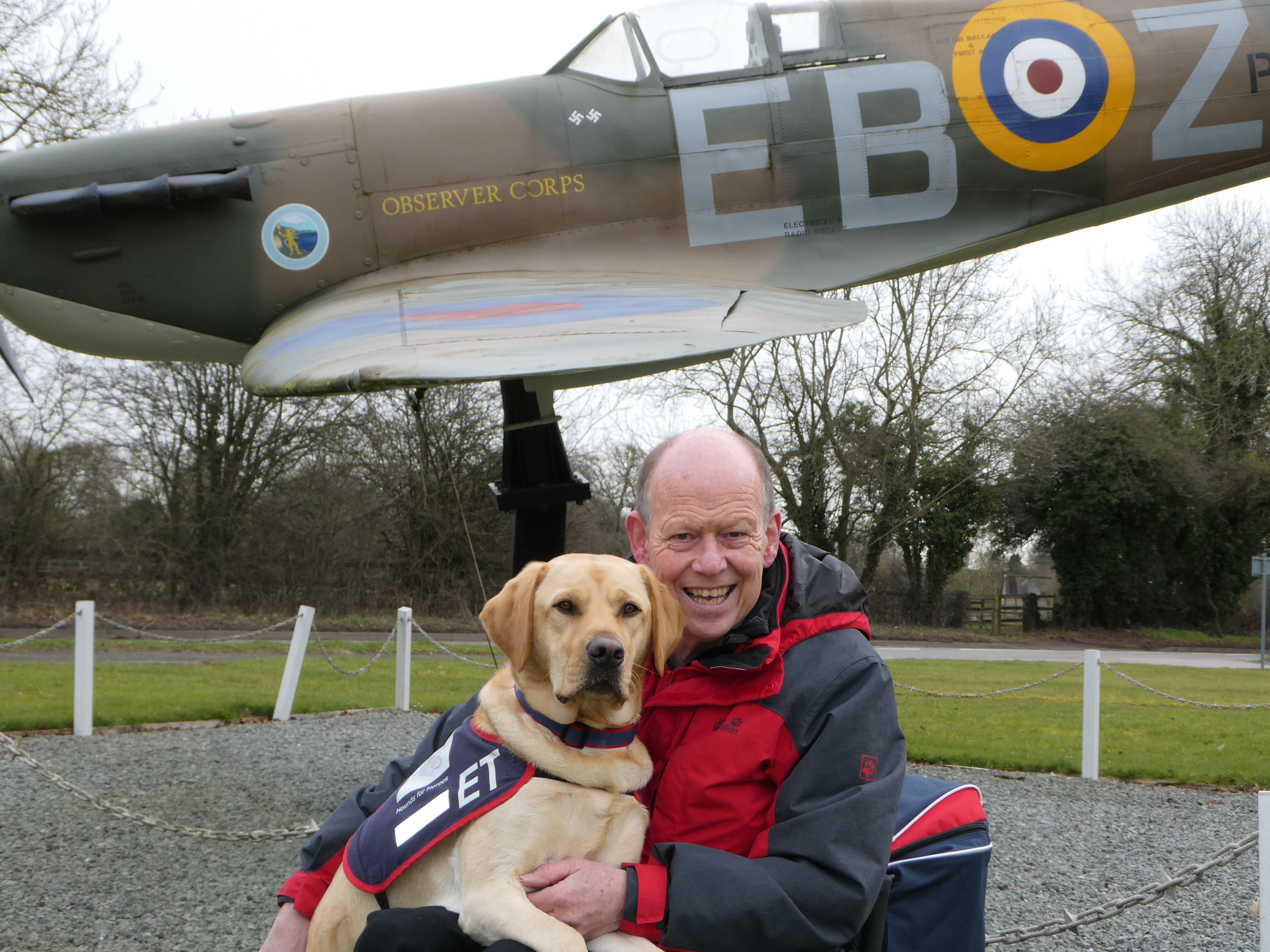 Image shows veteran with Assistant Dog in front of guardian gate Spitfire model. 
