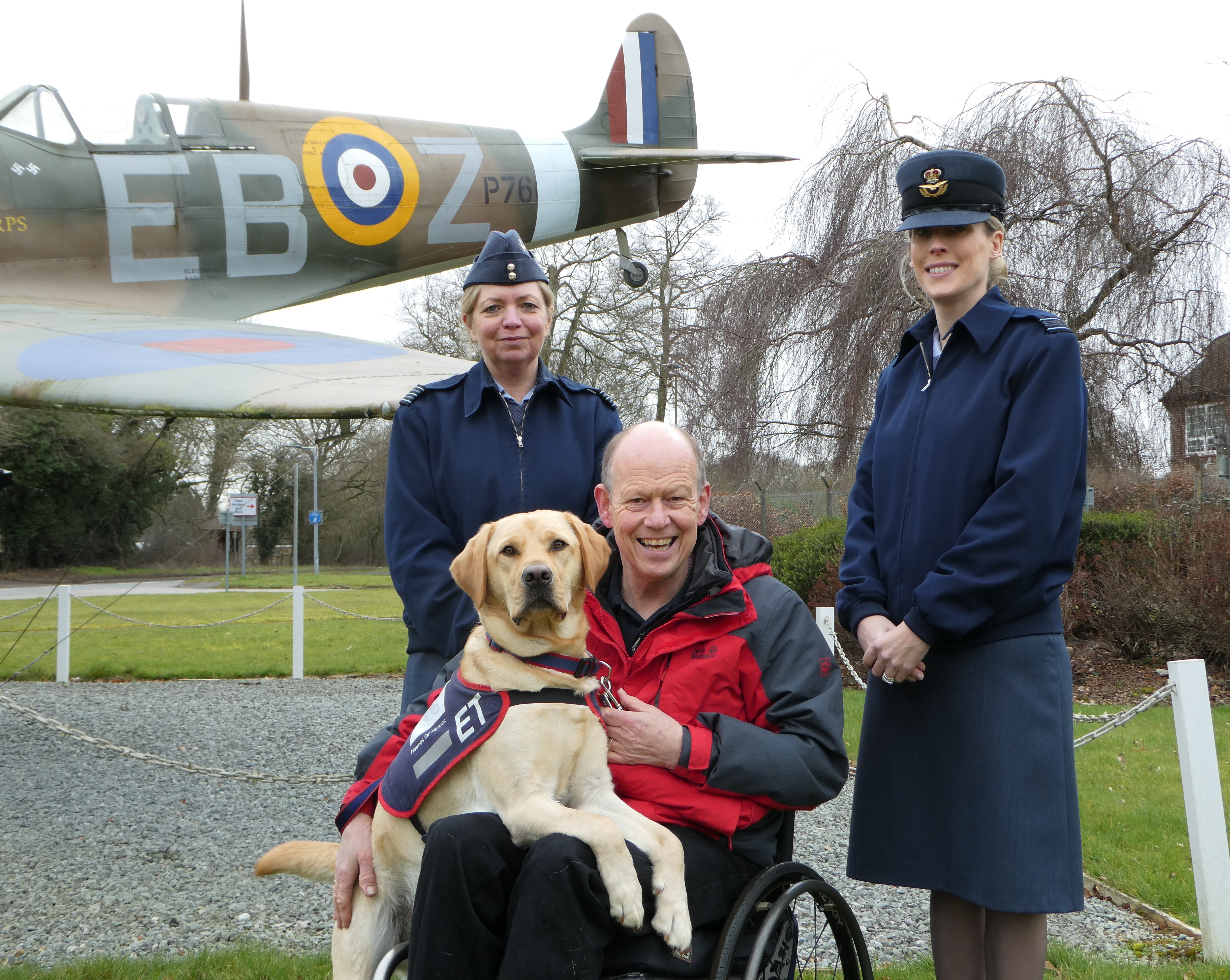 Image shows RAF Personnel and veteran with Assistant Dog in front of guardian gate Spitfire model. 