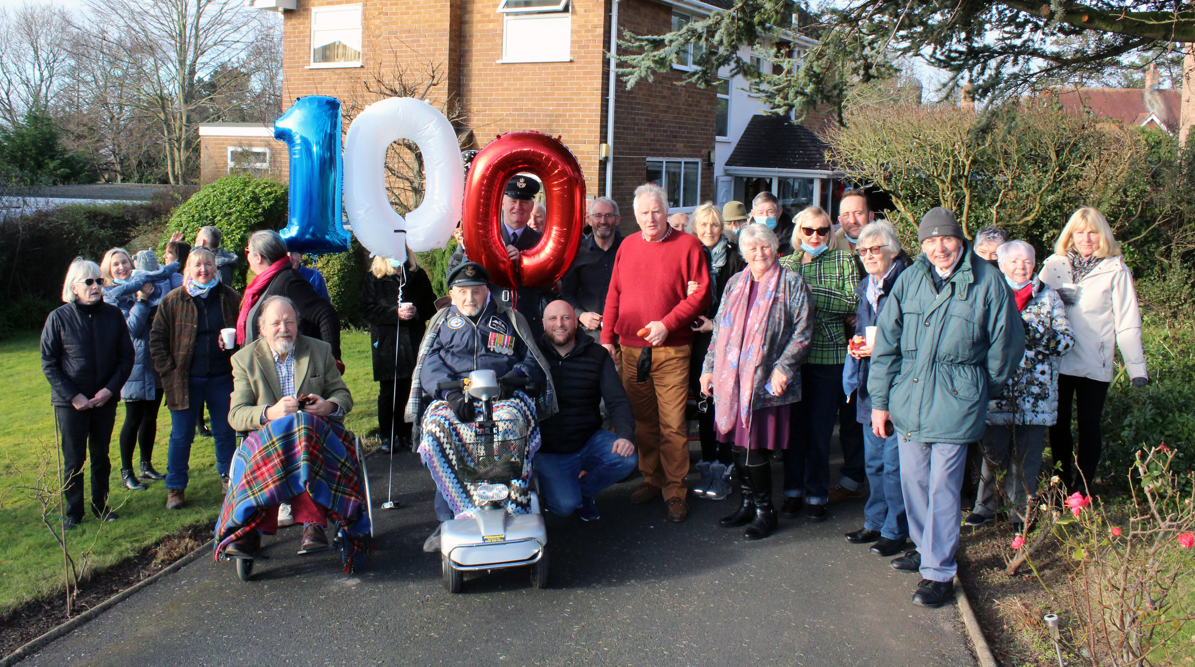 Eric sits in wheelchair outside, with family and personnel, and 100 shaped balloon.