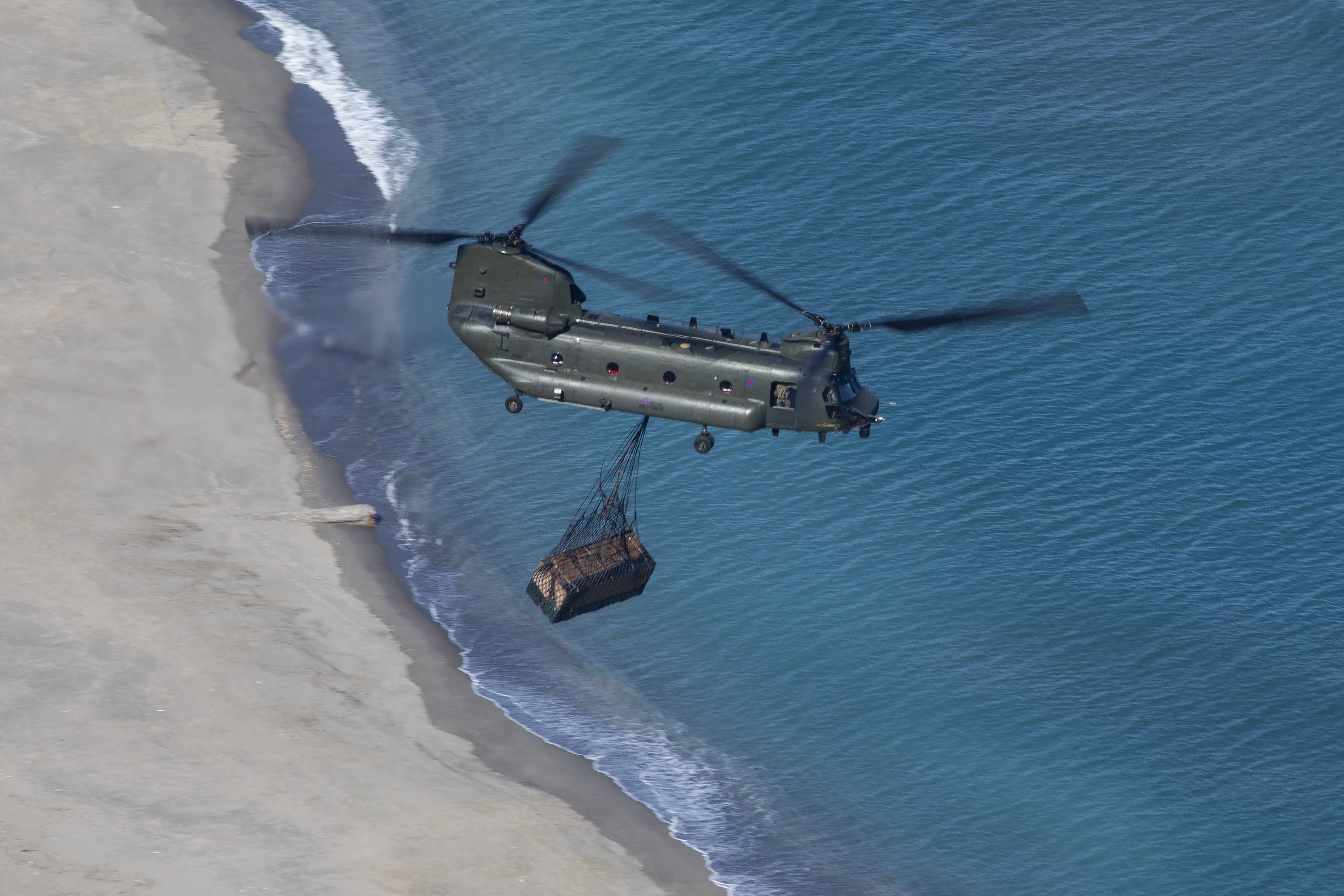 Chinook carries loaded sling of cargo over coastline.