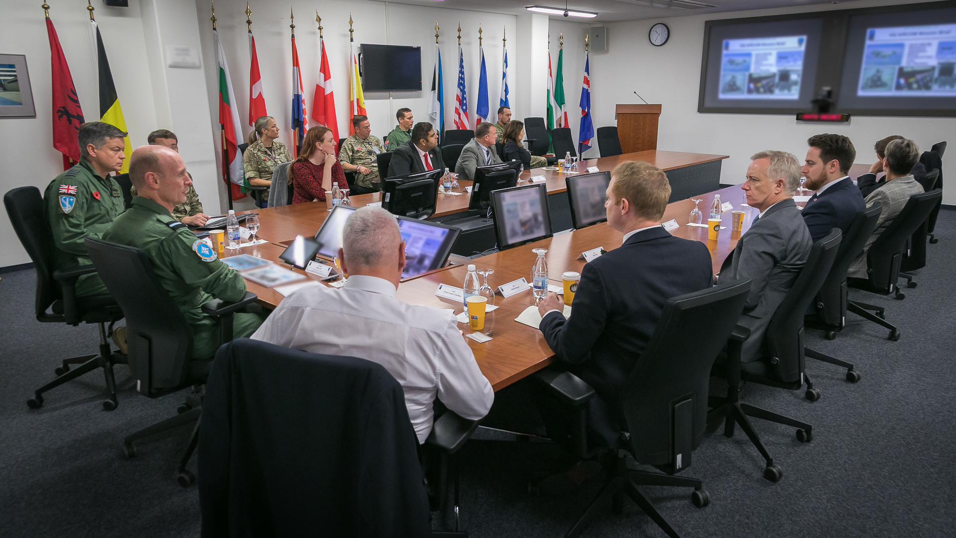 Personnel sit around conference table with flags, computers, and presentation board.