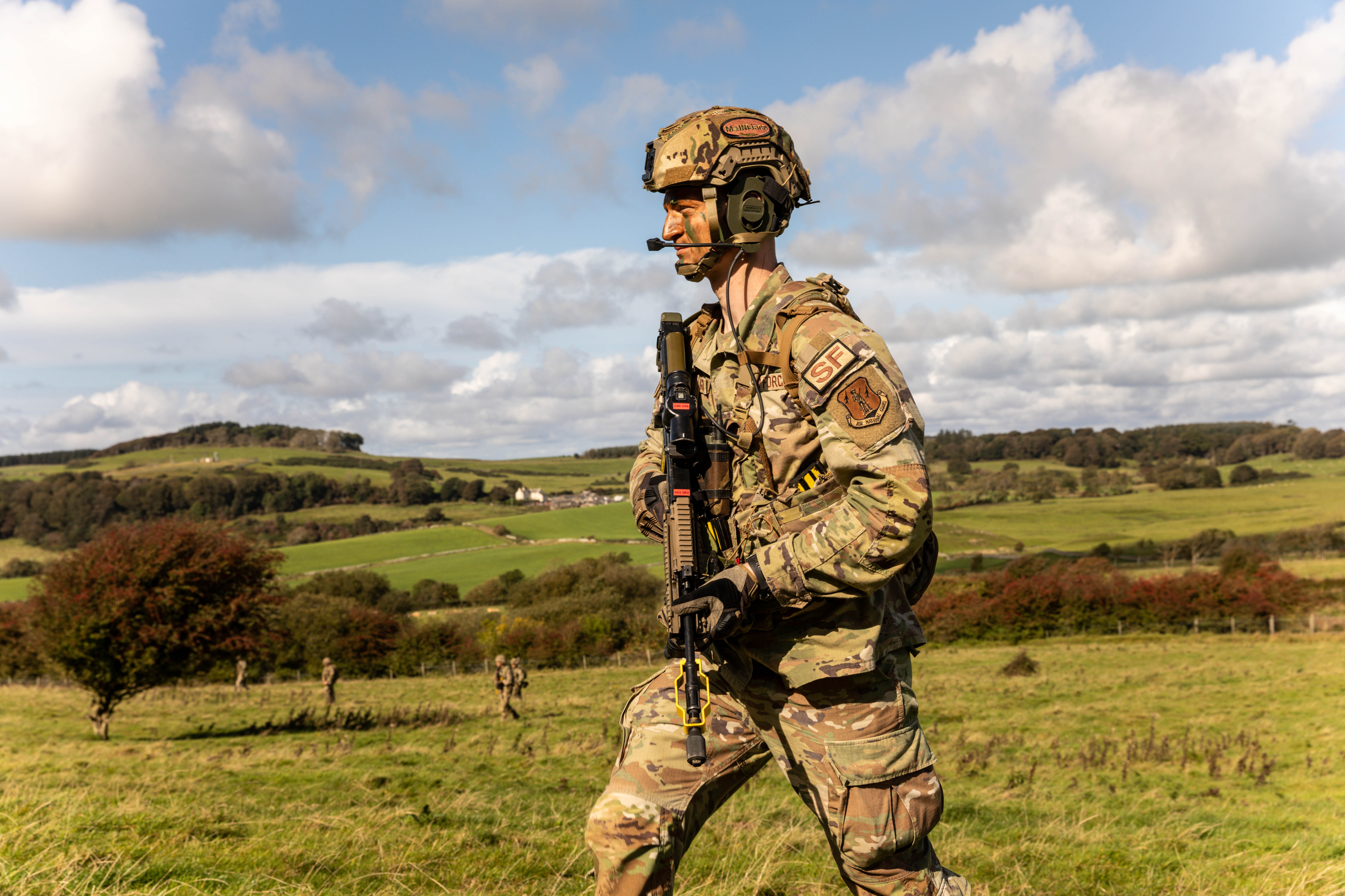 United States Air Force Reservist in combat uniform, carrying rifle against backdrop of Scottish countryside.
