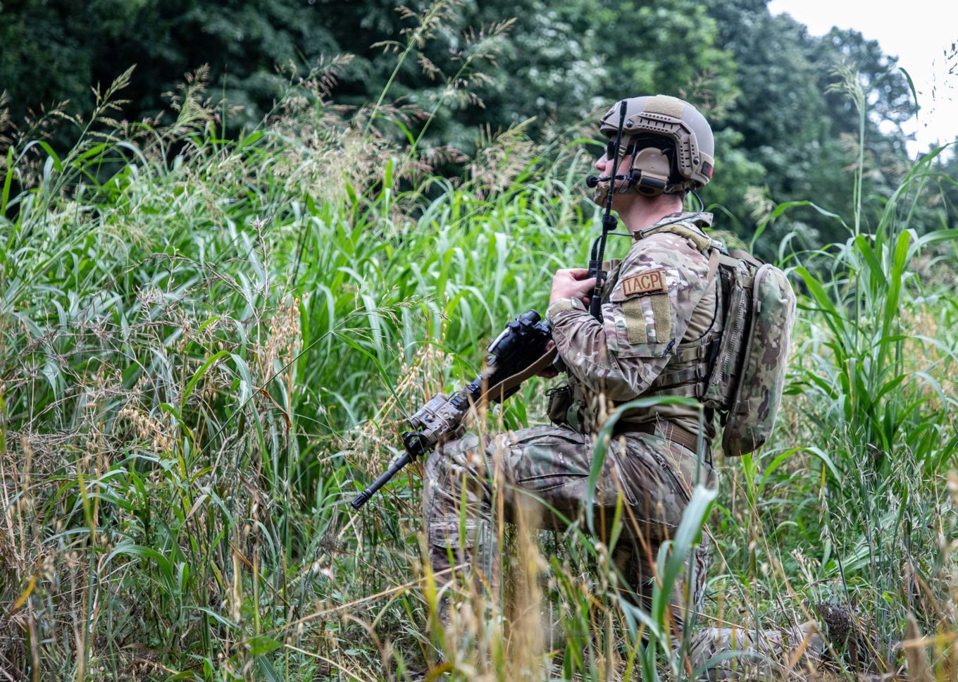 Personnel holds rifle and radio amongst the tall grass. 
