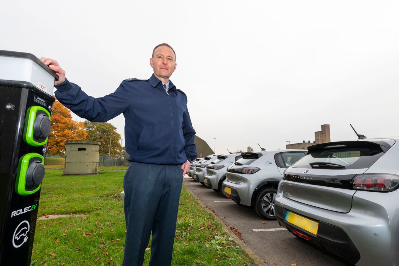 Image shows the Warrant Officer standing in front of the six new Peugeot e-208 electric vehicles at RAF Leeming while leaning on one of the new charging points.