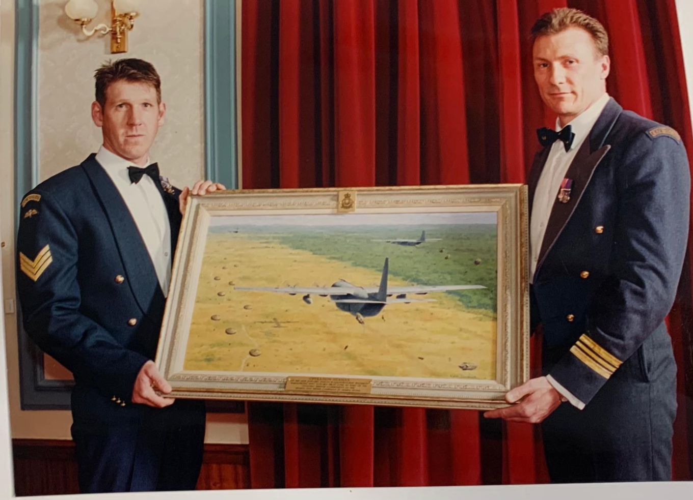 Image shows two members of II Squadron RAF Regiment holding the oil painting depicting the parachute drop.