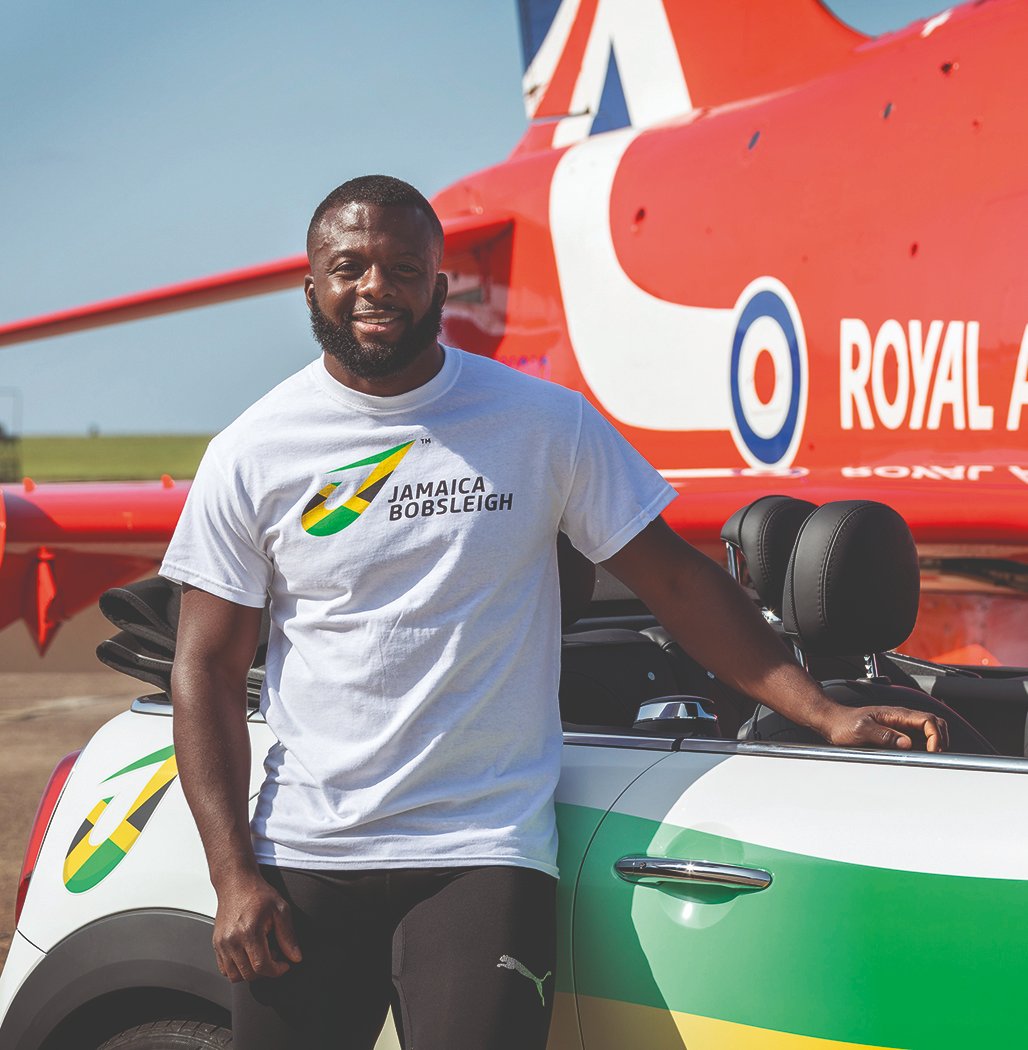 Shanwayne stands by Mini car and rear end of an aircraft.