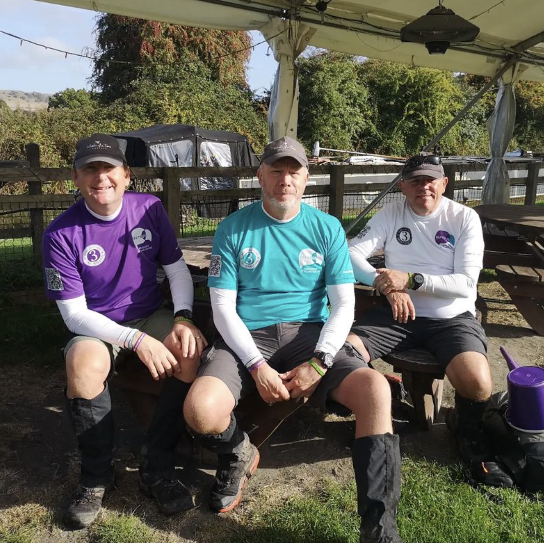 Image shows three dads sitting on a table bench under a canopy. 