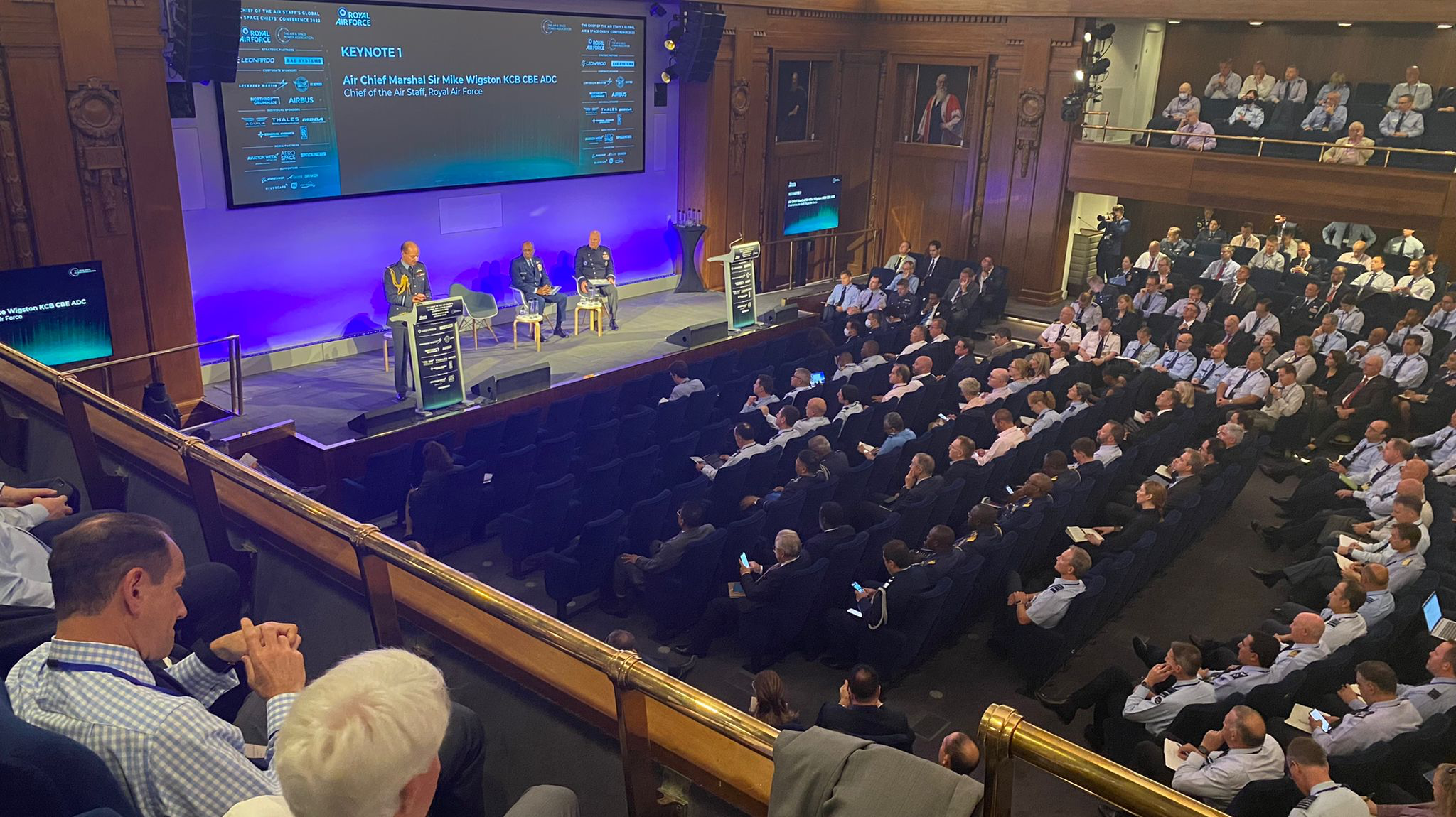 Image shows audience listening in conference room, with presentation screen and stage.