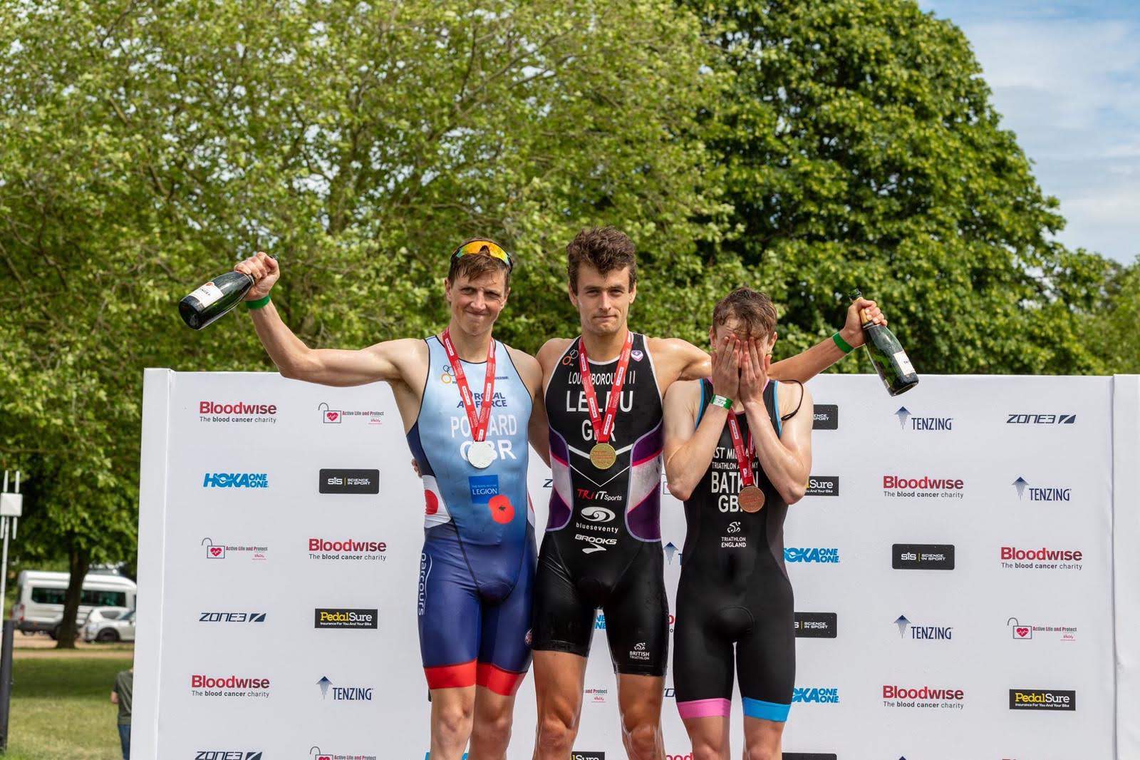 Image shows RAF aviator standing on podium with two other medal winners and champagne bottles.