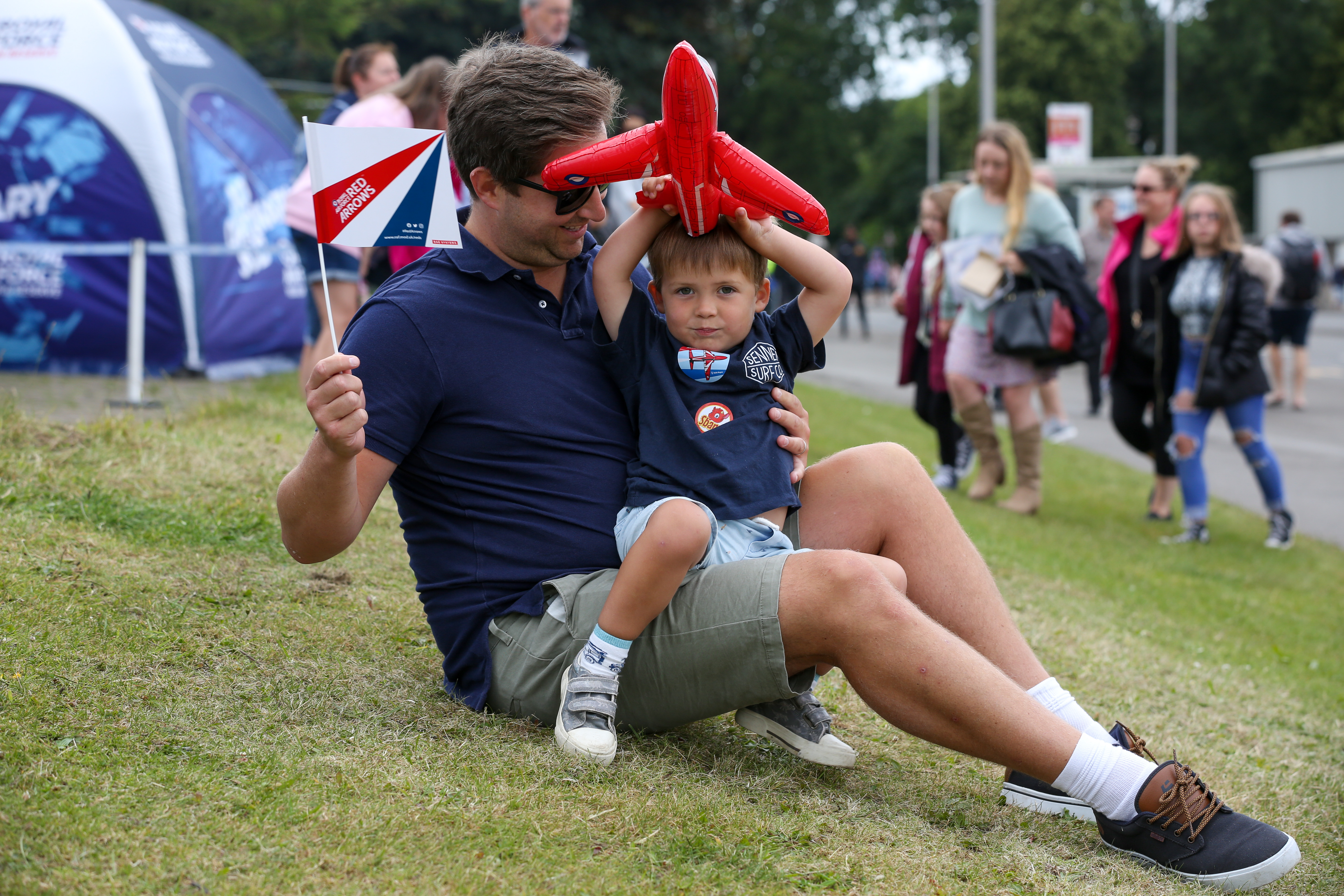 Image shows a child sits on father while holding a blow up aircraft.