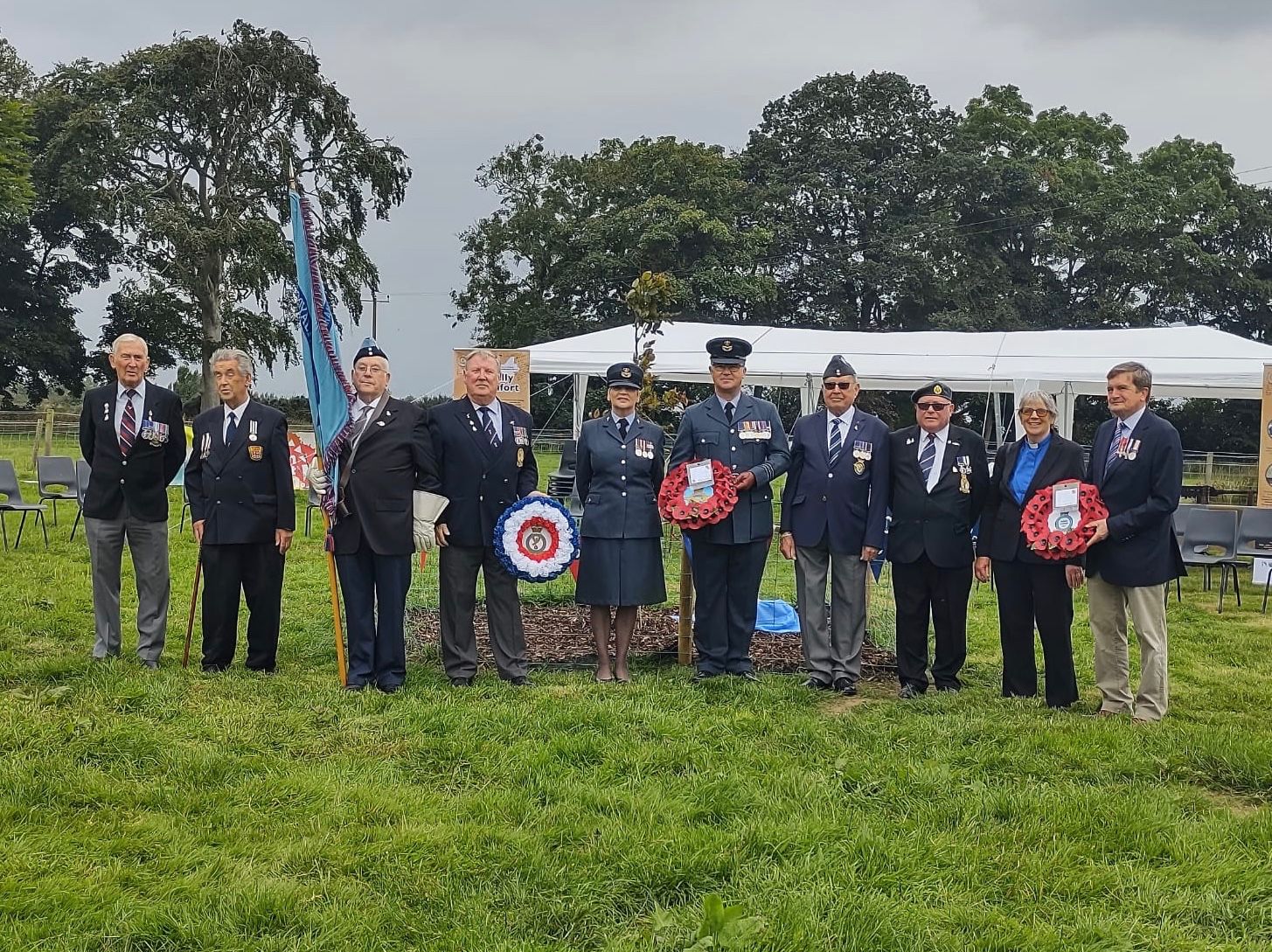 Personnel stand outside marque with poppy wreaths and Flag Colour. 