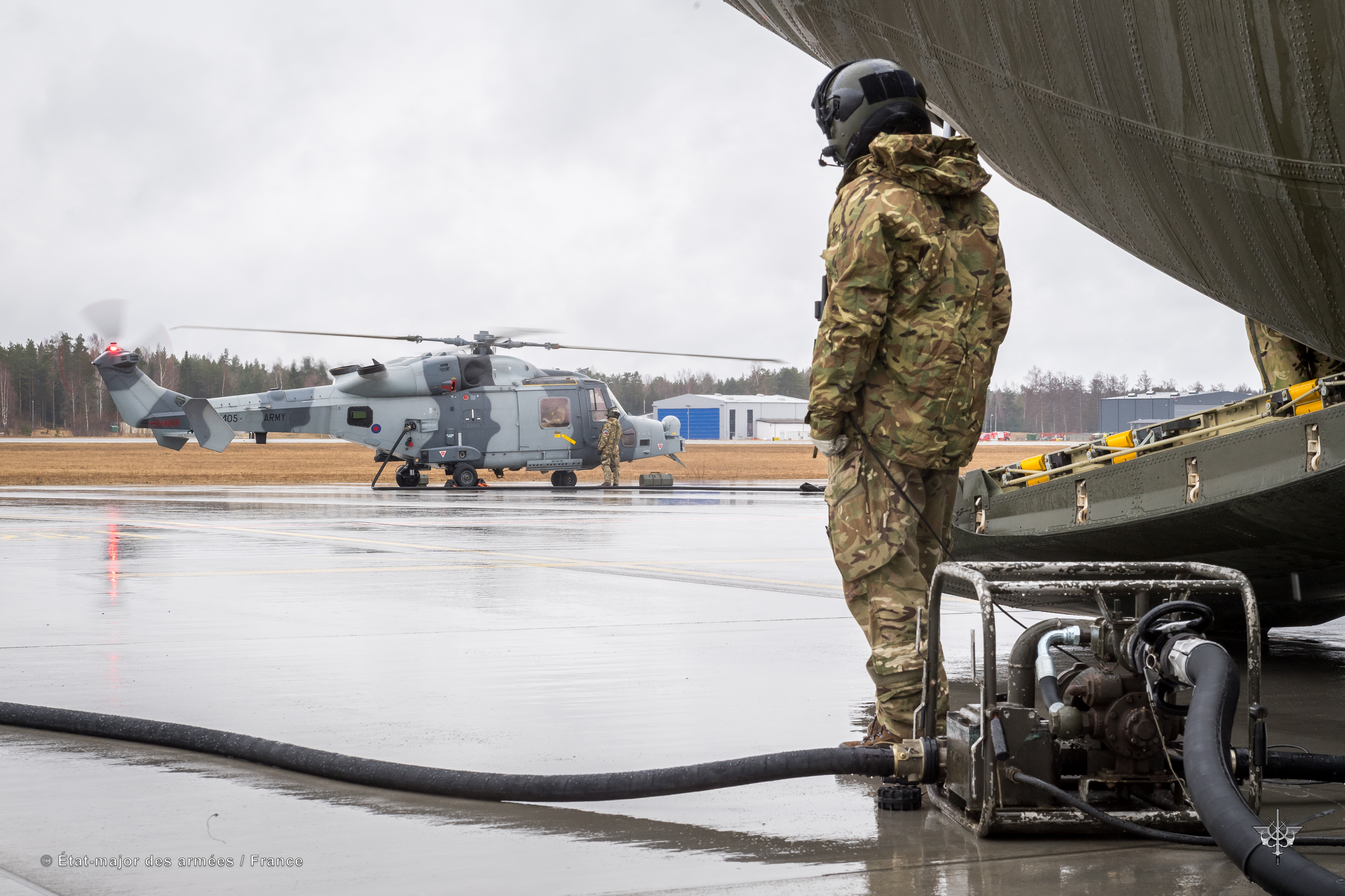 Pilot stands by refuelling pump, looking towards helicopter on airfield.