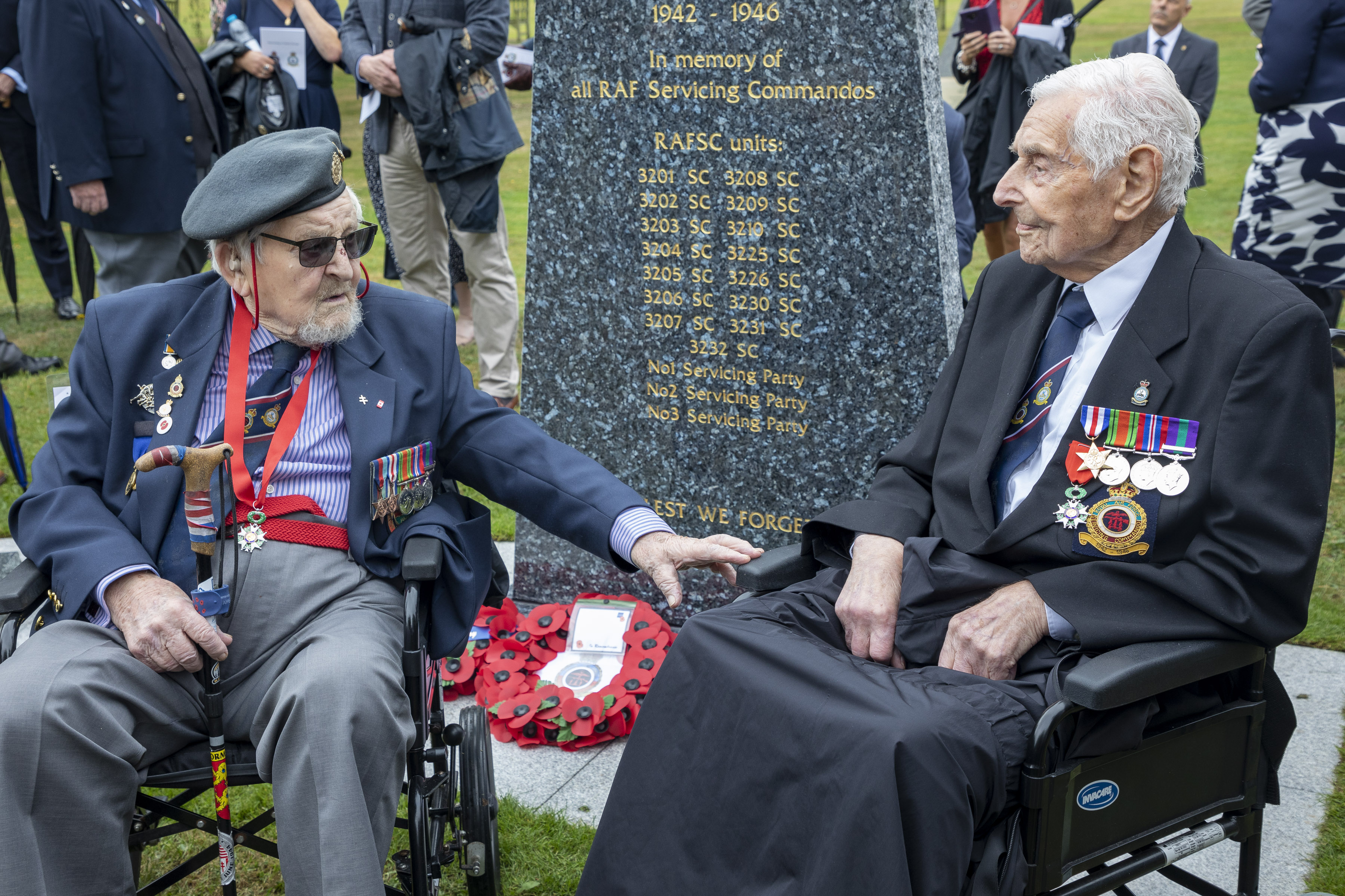 Two veterans in wheelchairs sit by memorial stone.