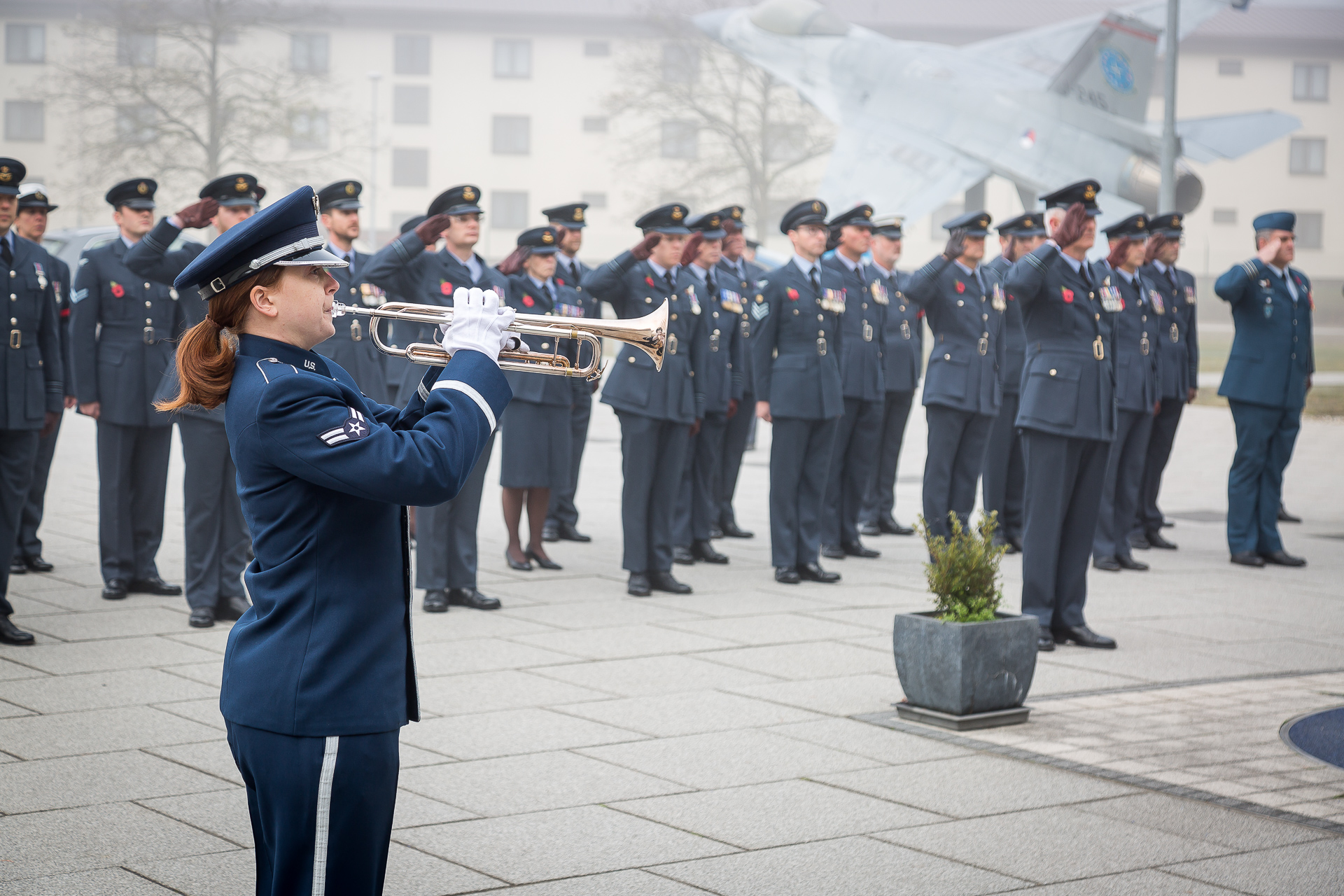 Corporal Hogan plays the bugle while personnel in parade salute. Model aircraft in the background.