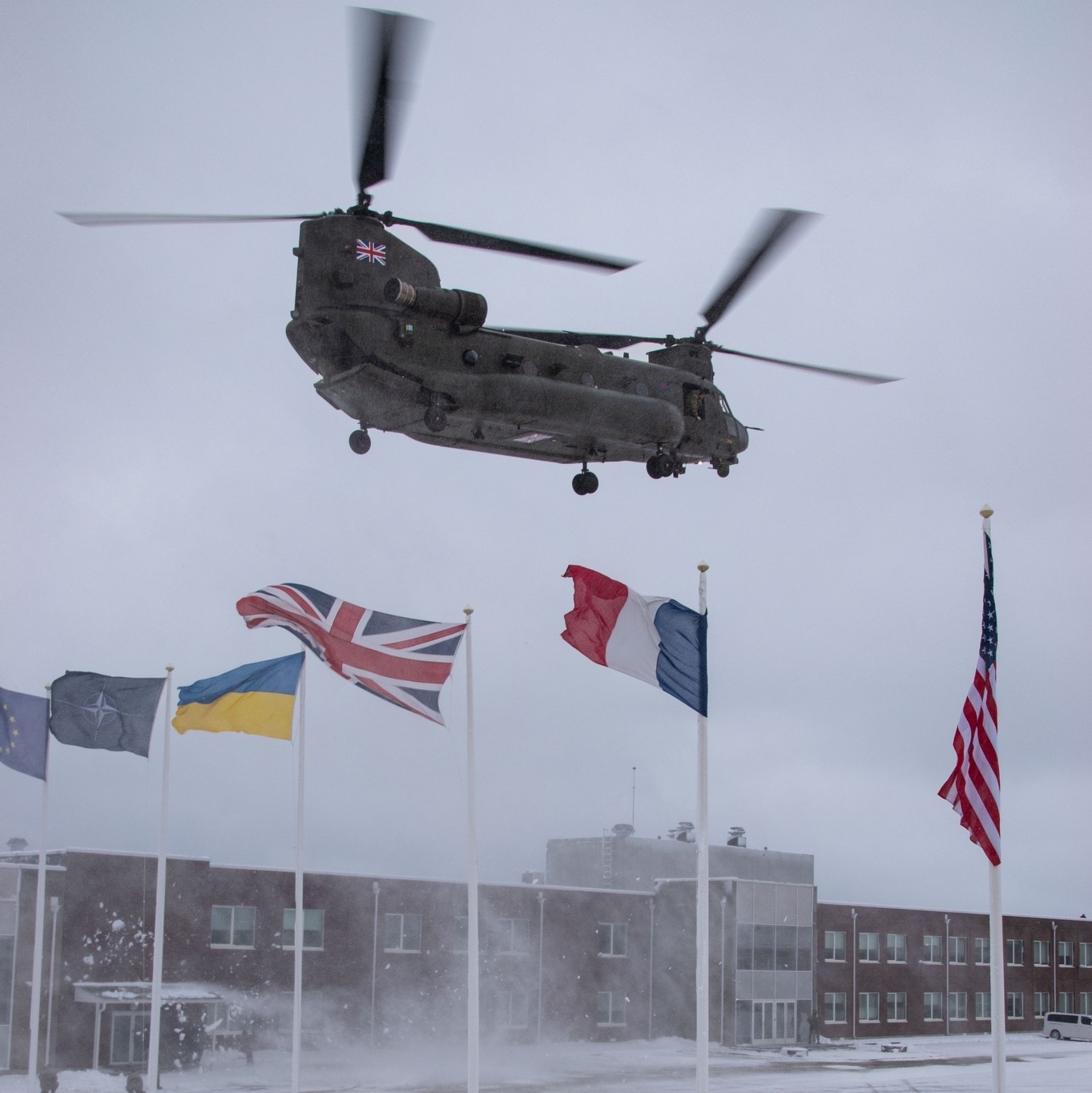 Image shows RAF Chinook flying over a snowy nation ground with flags and building headquarters.