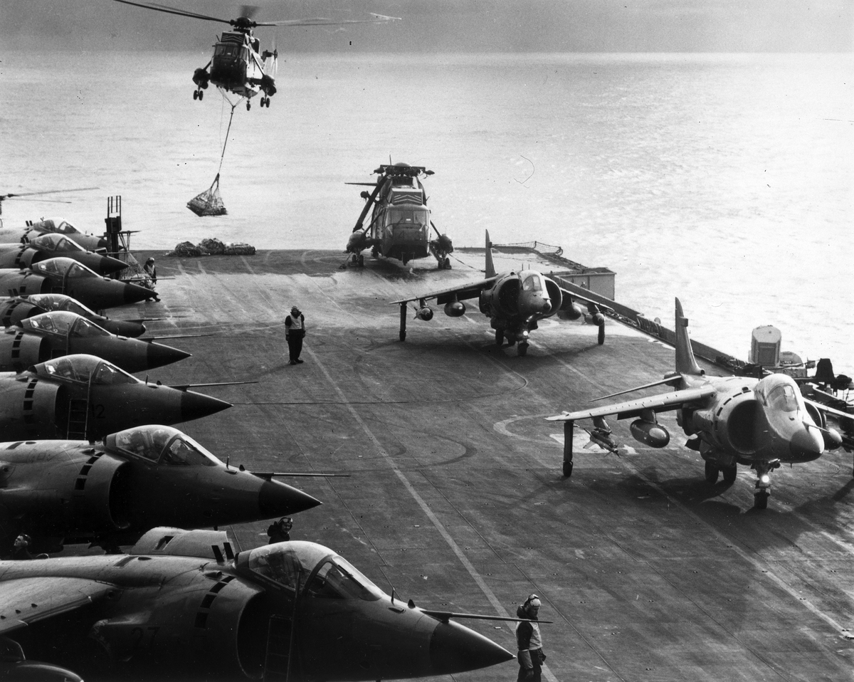 Aged image of helicopter carries sling load to deck of HMS ship, with aircraft on the taxi way..