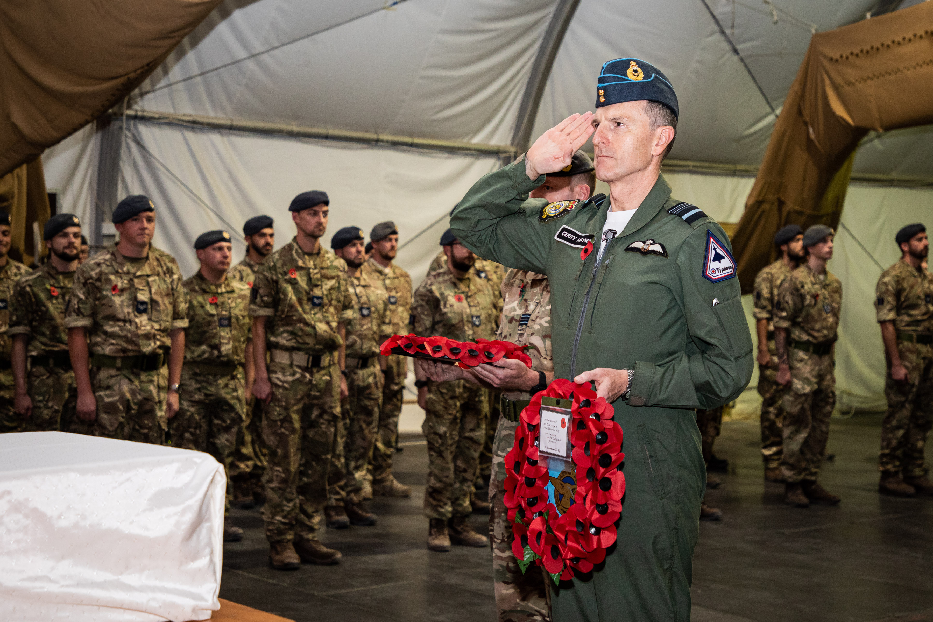 Personnel stand inside tent, with Air Marshal Mayhew saluting as he lays poppy wreaths.
