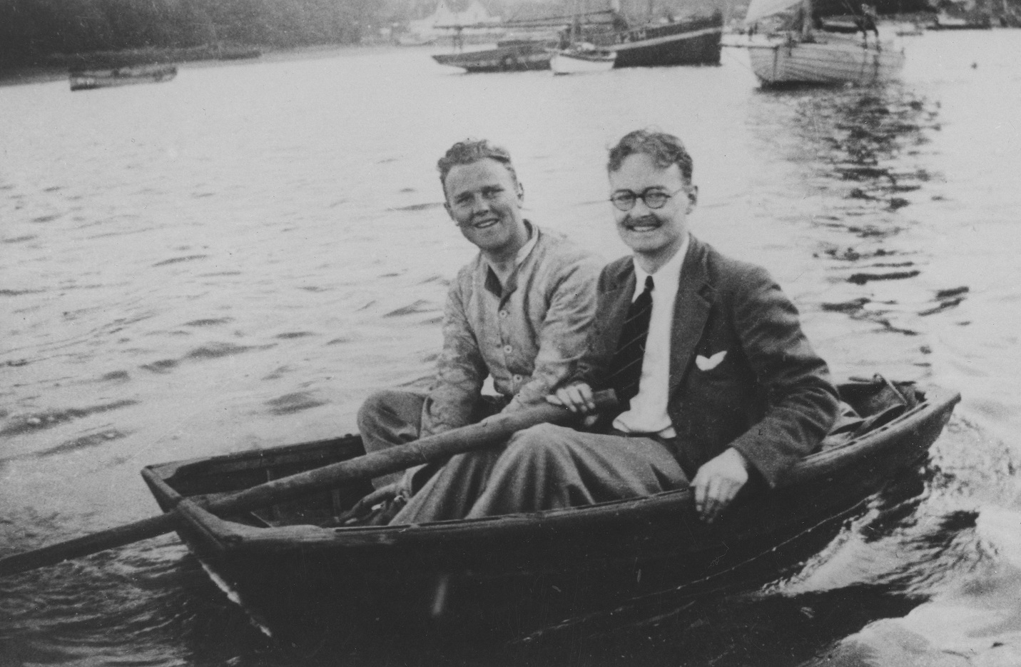 Black and white image shows two men sitting in a small rowboat on the river.