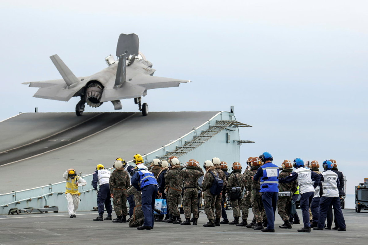 Lightning takes off from aircraft carrier, with ground grew gathered nearby.