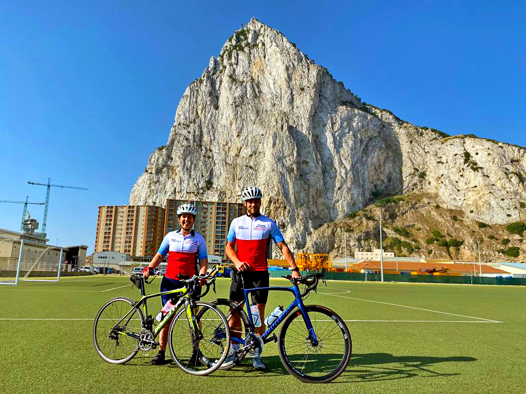 Riders stand with bikes, on the tennis courts by large Rock of Gibraltar.