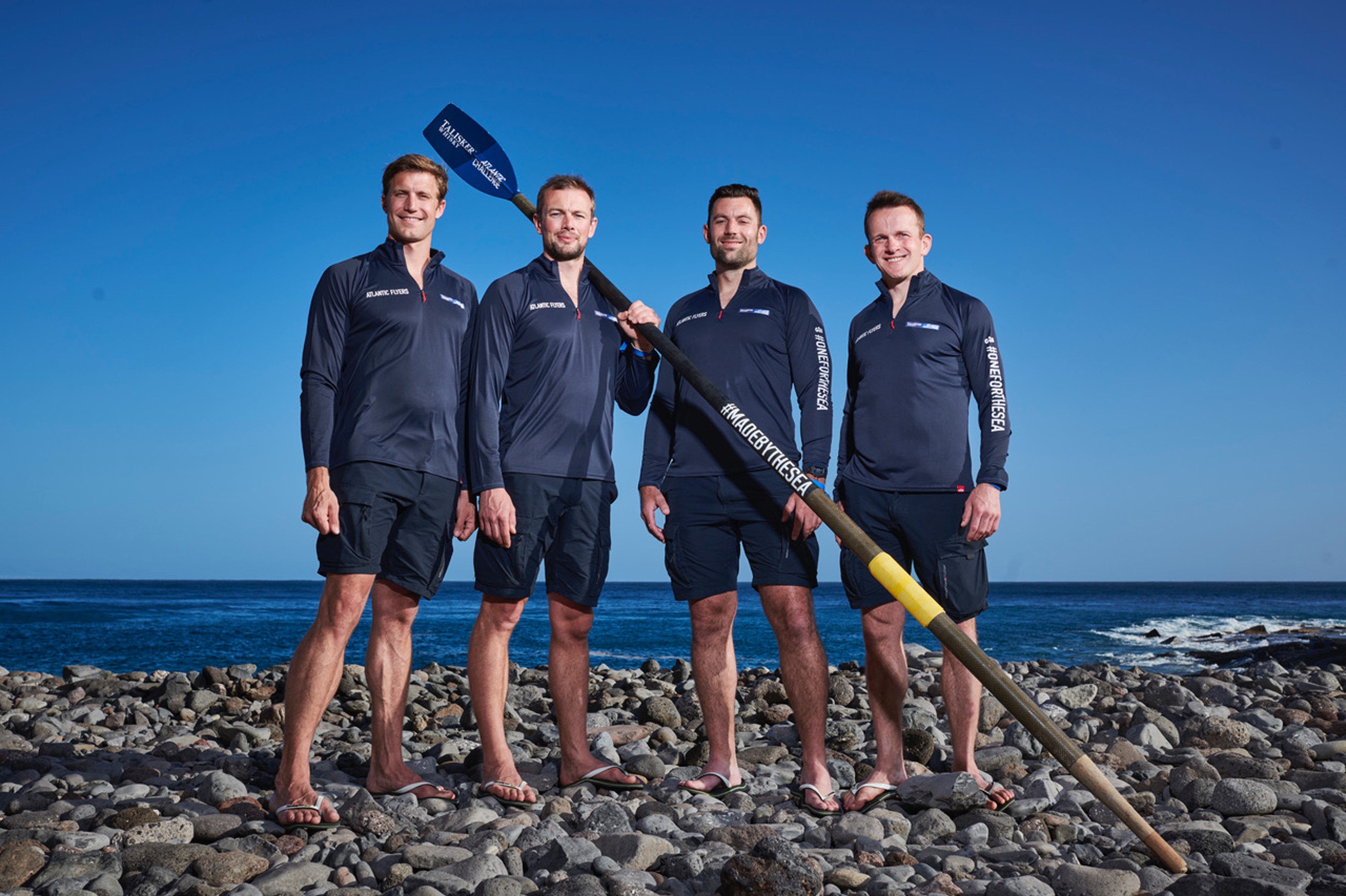 Rowers stand on rock beach with oar.