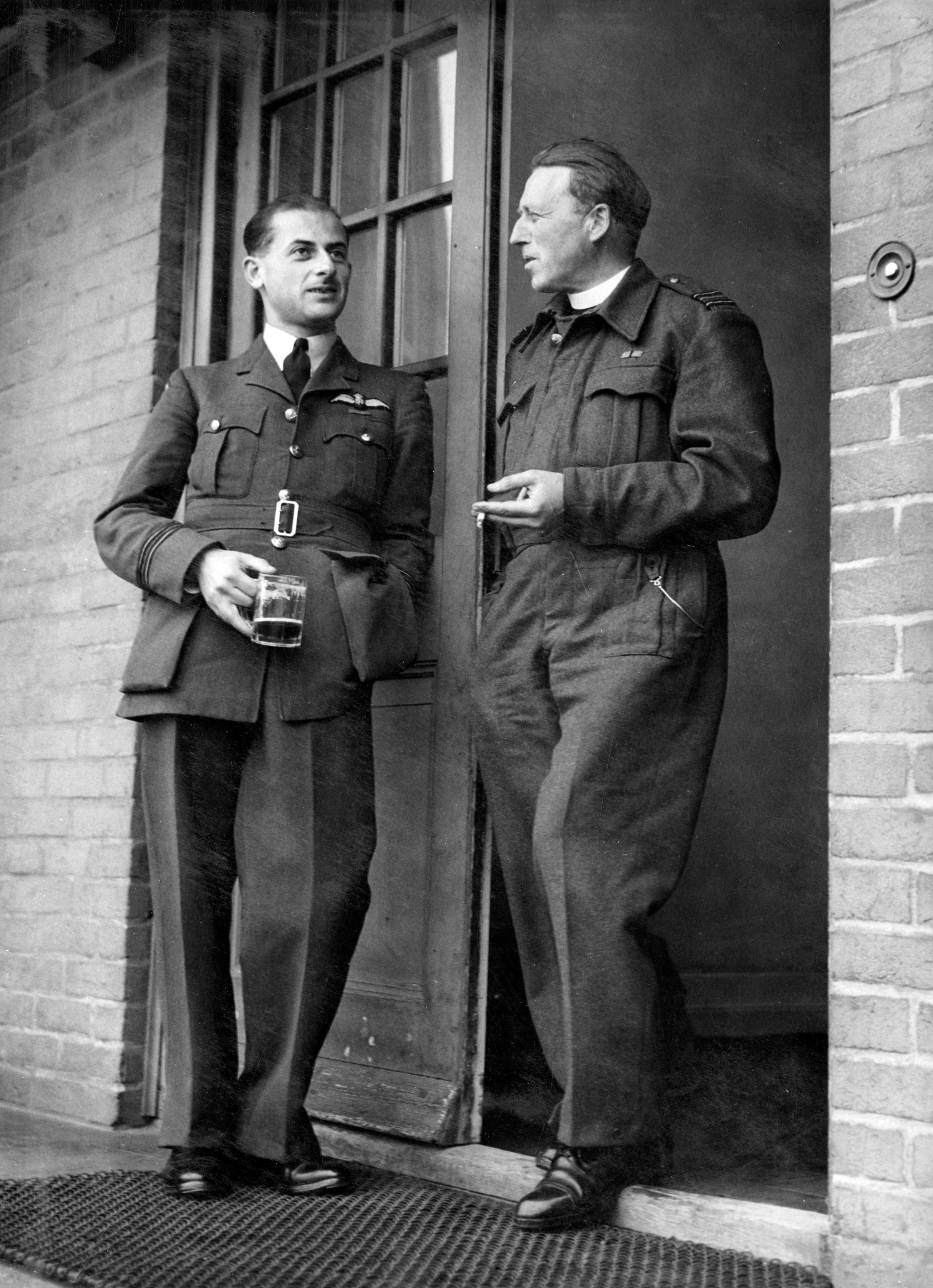 Flight Lieutenant and chaplain talk at the door; one smokes while the other drinks.