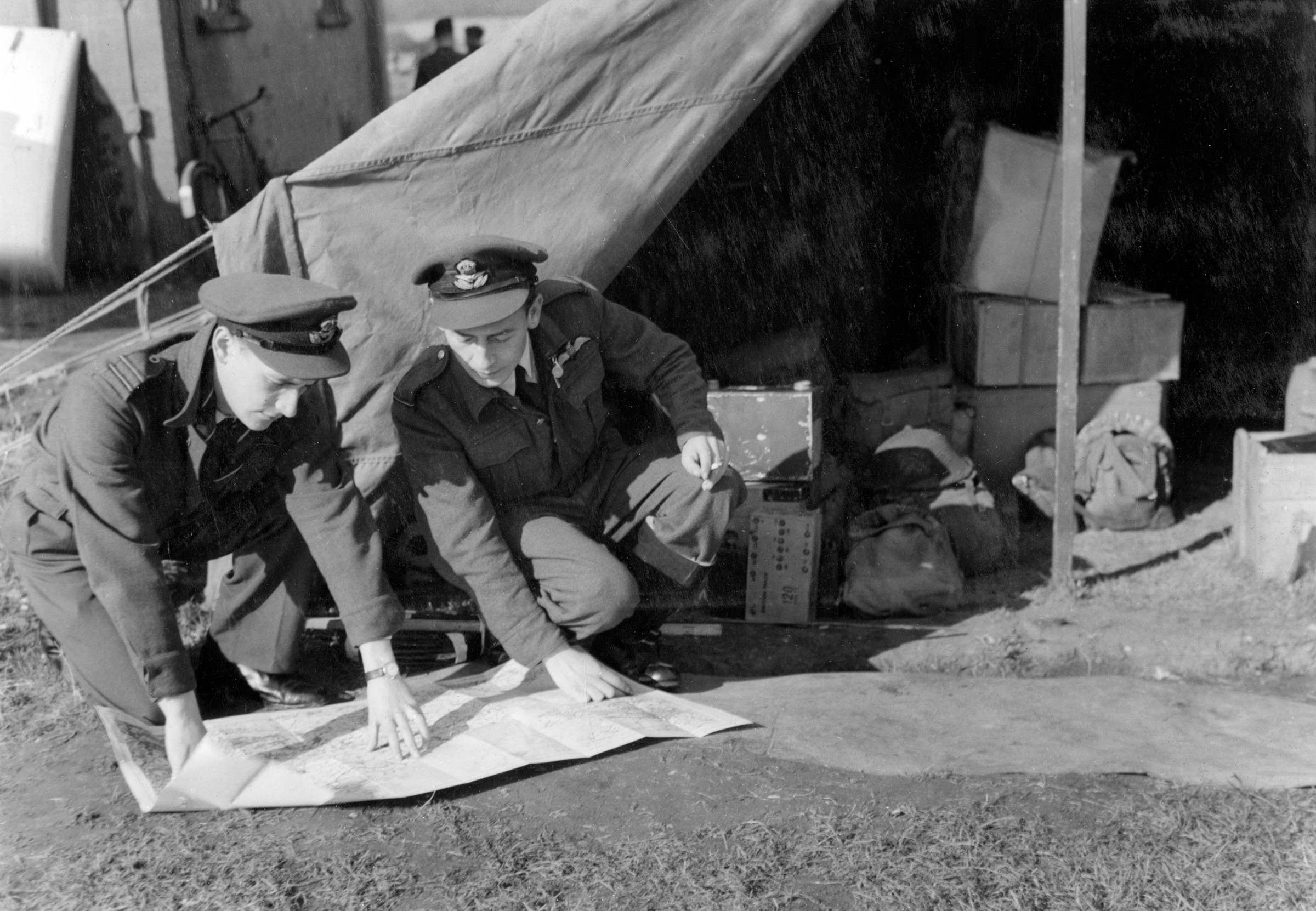 Personnel study a map on the ground, in front of a tent on the airfield.