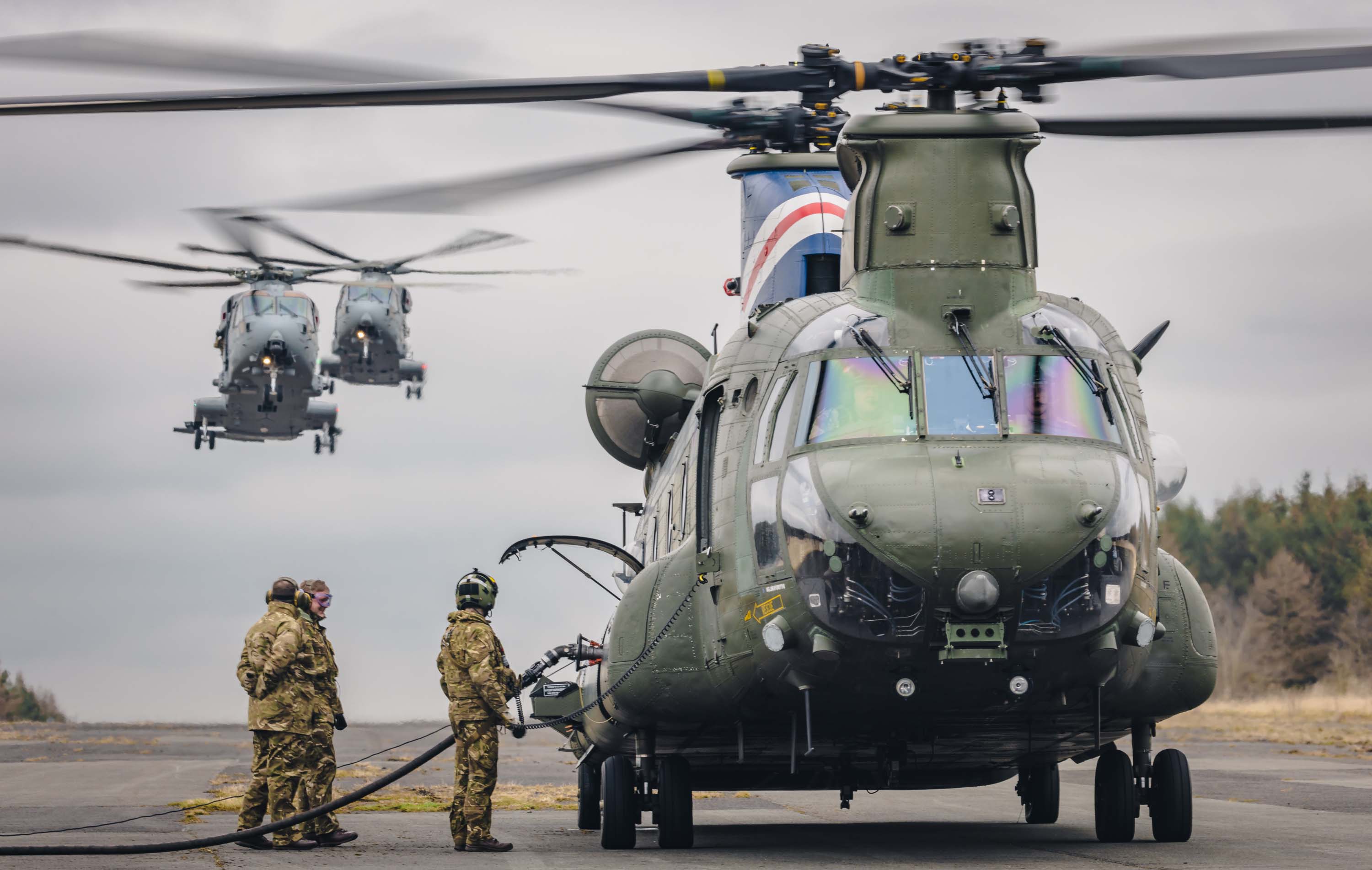 Image shows RAF aviators refuelling a helicopter on the airfield while two more come into land.