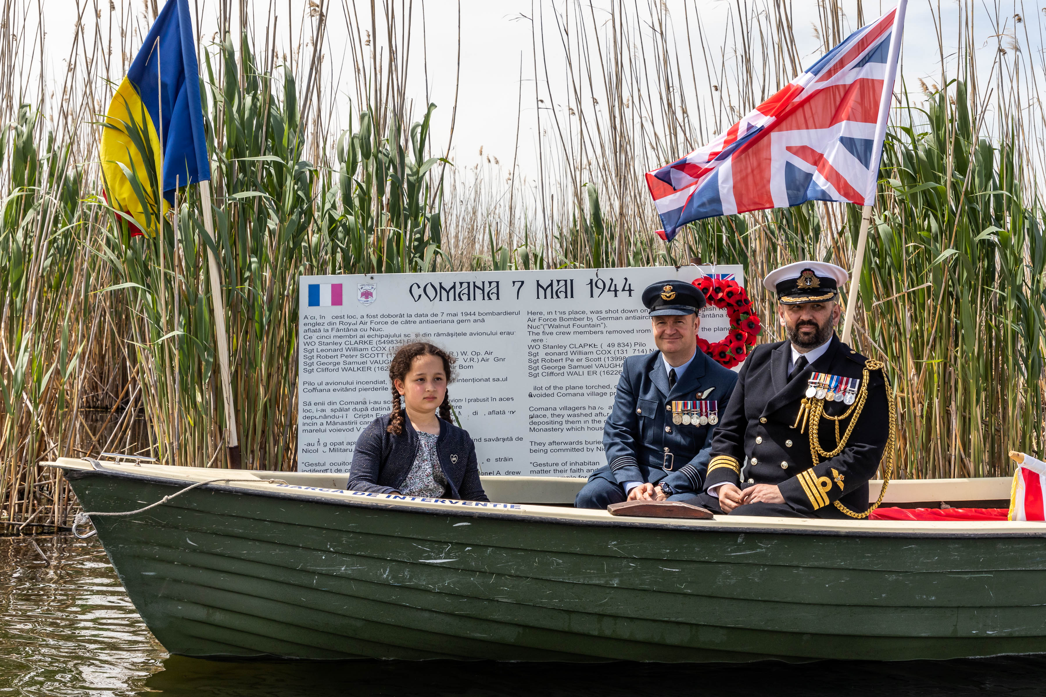Personnel in sailboat among river reeds and flags.