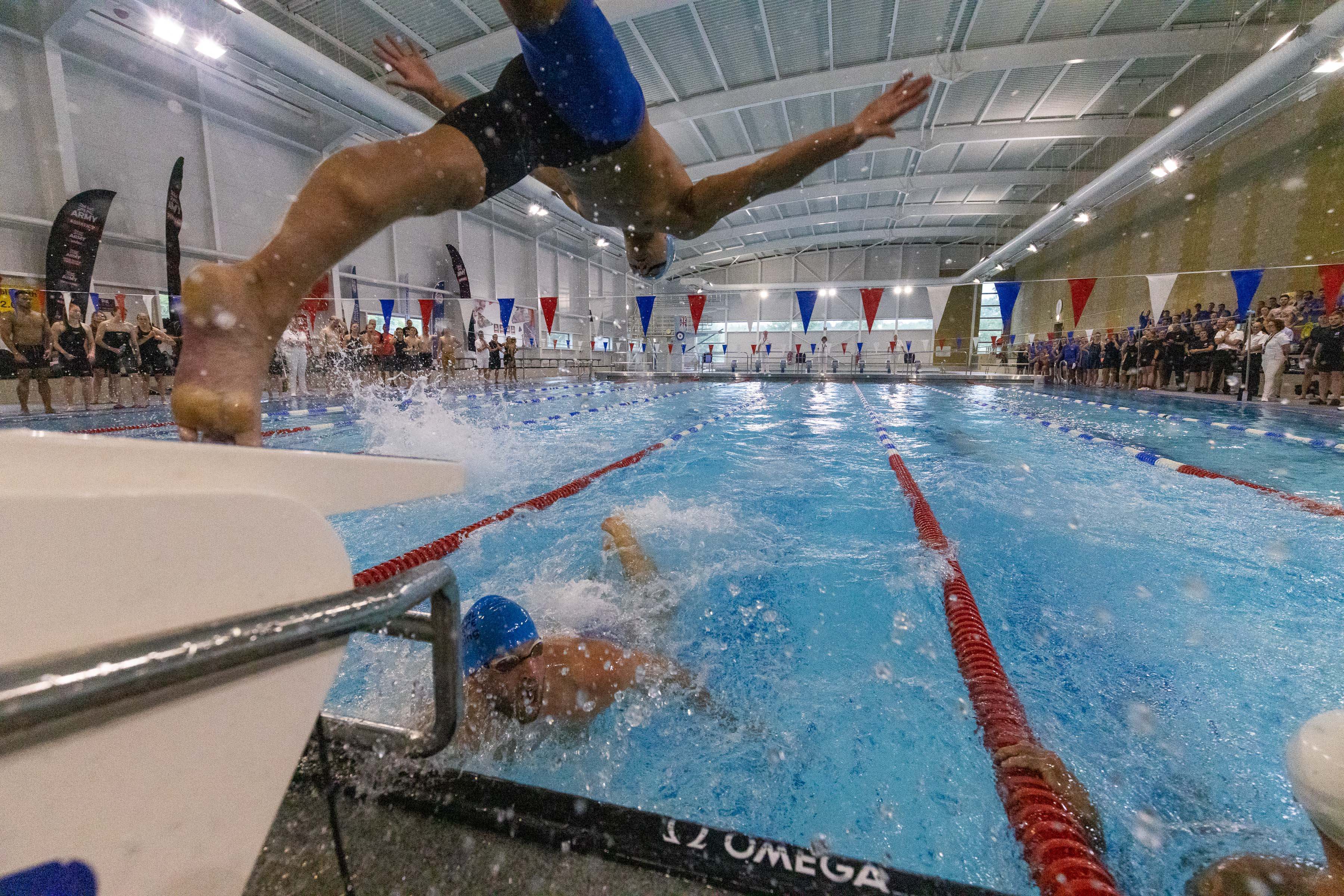 Poolside shot of 1 swimmer reaching the side and the next diving in.