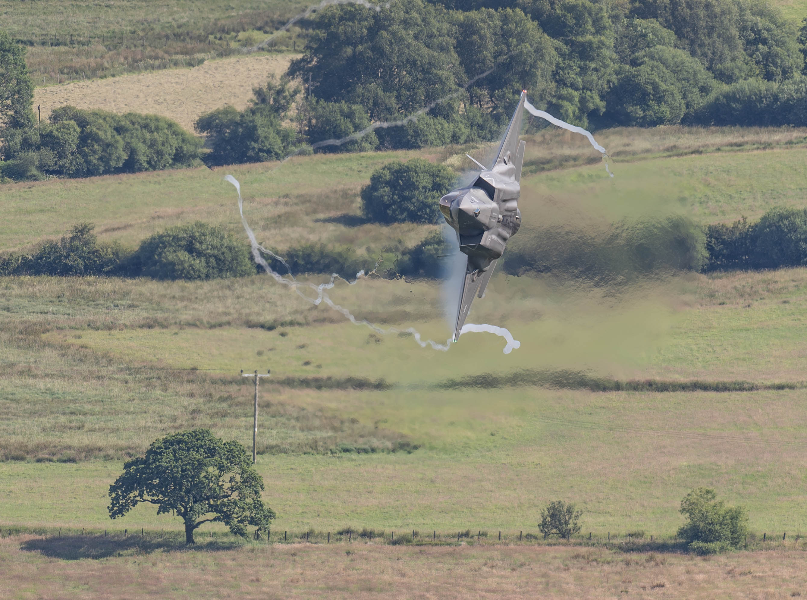 "F-35". An F-35B in low flight over the countryside, fully banked to its right so that the right wing is perpendicular to the ground.