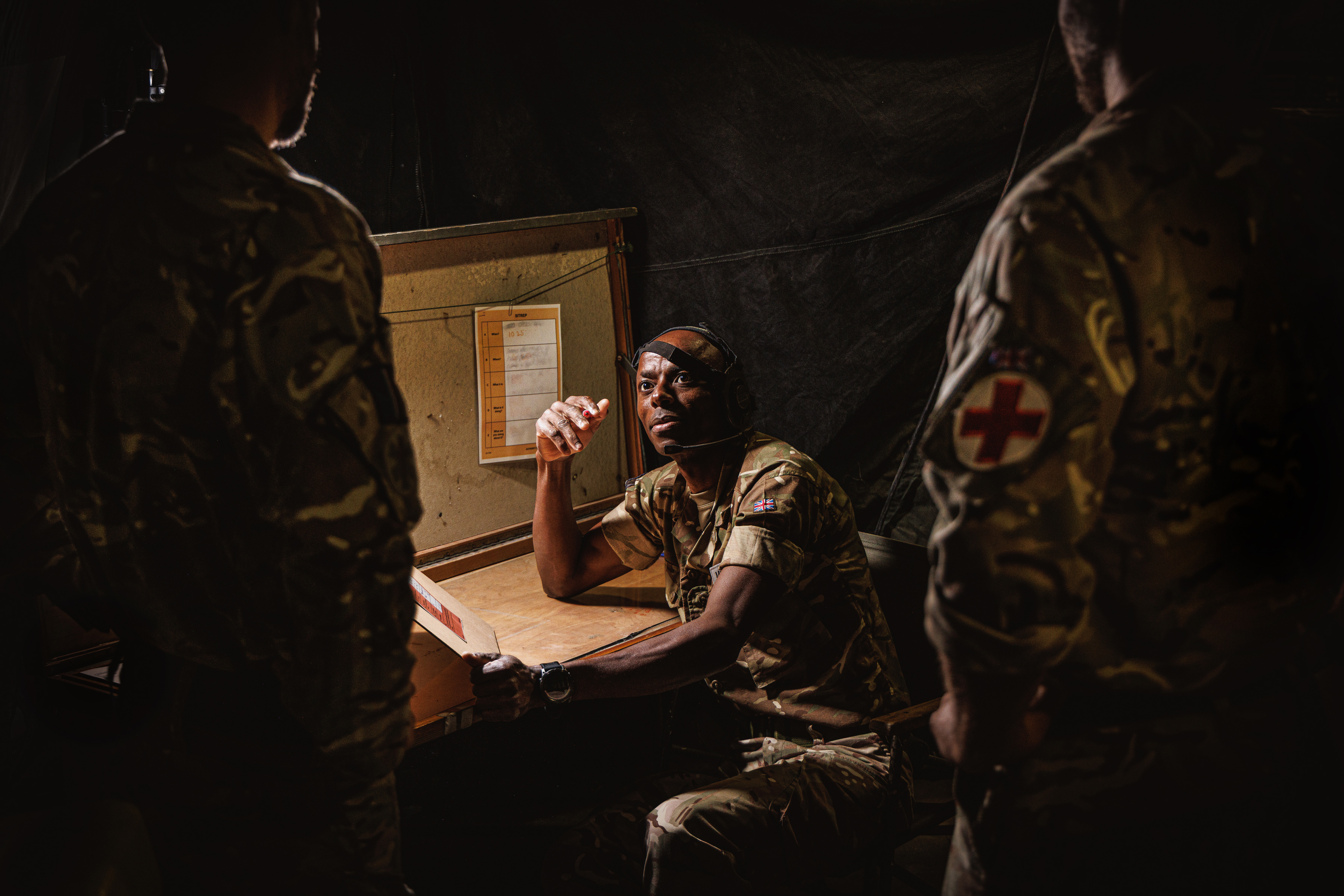 "Medical Advice". Three RAF Medical personnel in a dark tent. One of them, caught by the light, faces the camera and is sat with a clipboard in their hand.