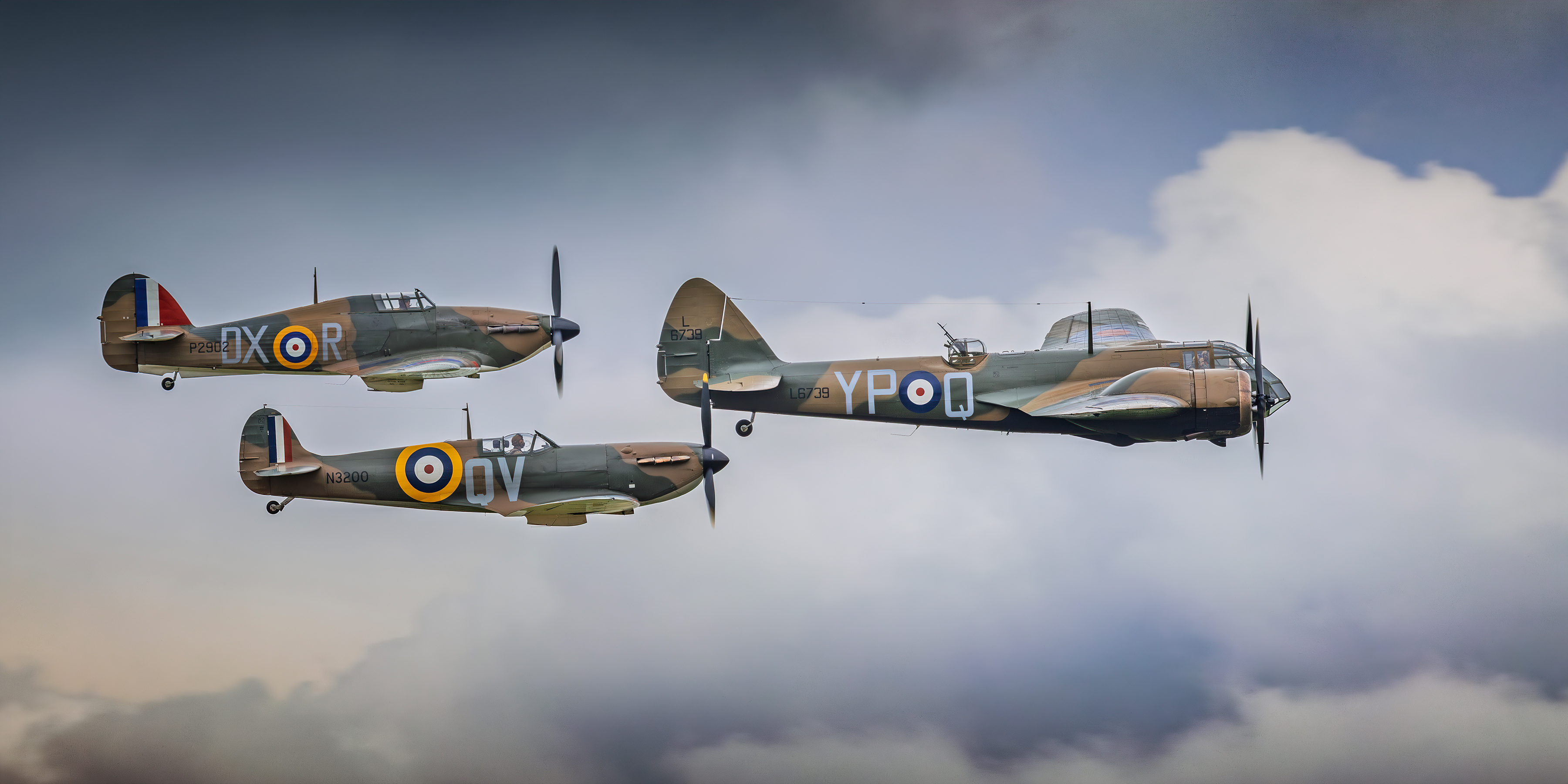 "Old But Gold". Three RAF Heritage aircraft fly in formation through cloudy skies. A Lancaster aircraft flies ahead of a Hurricane and a Spitfire.