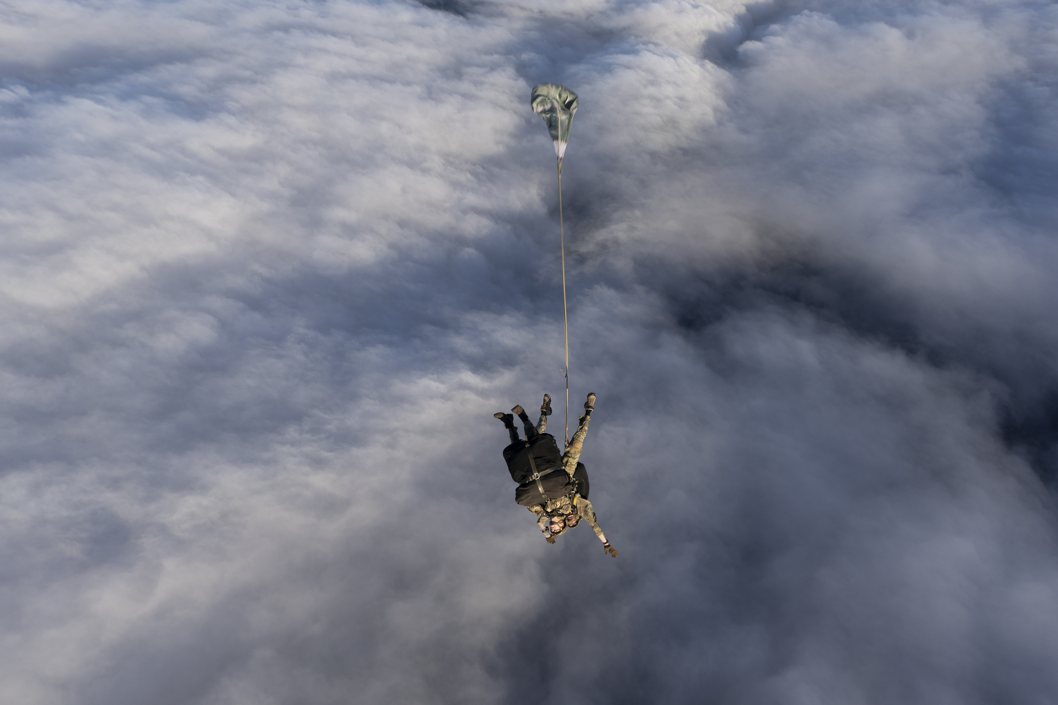 Tandem parachutists in military gear exit an aircraft above a thick layer of clouds, arms outstretched during freefall.