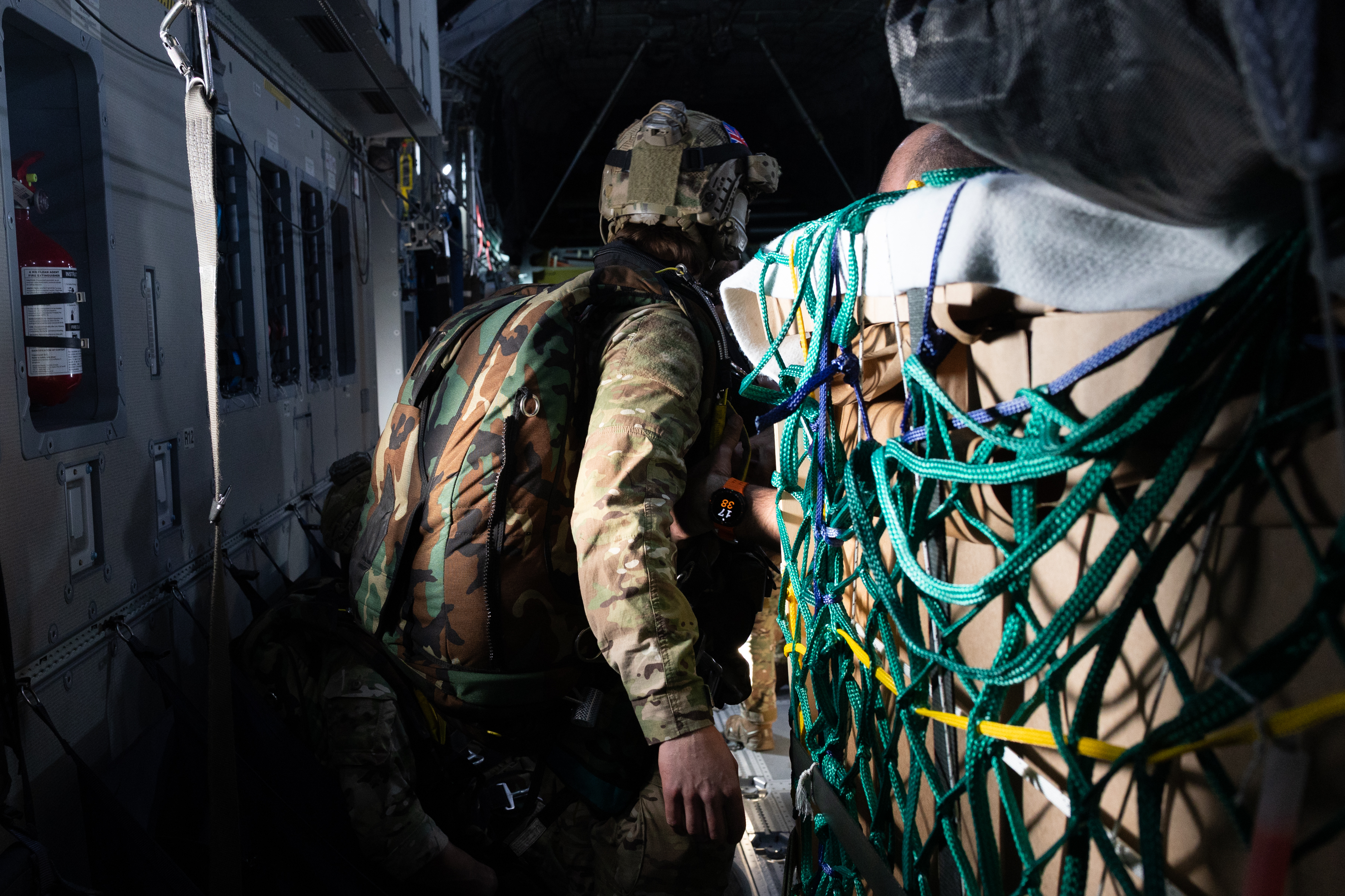 Close-up inside a cargo aircraft of a military crew member wearing a helmet and parachute pack, standing beside tightly netted cargo during loading or preparation.