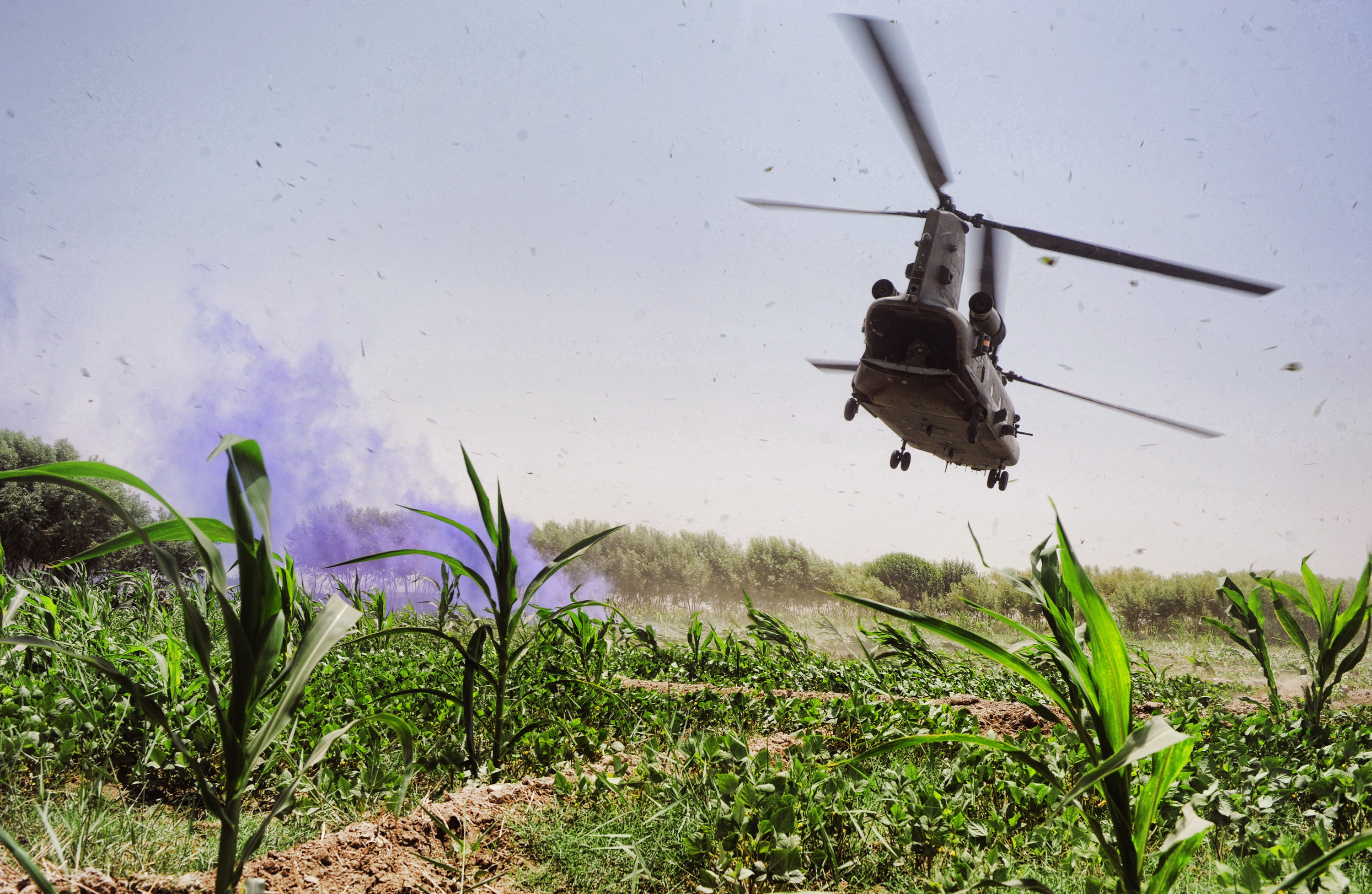 An RAF Chinook flying low-level over a field in Afghanistan with purple smoke marking a location.