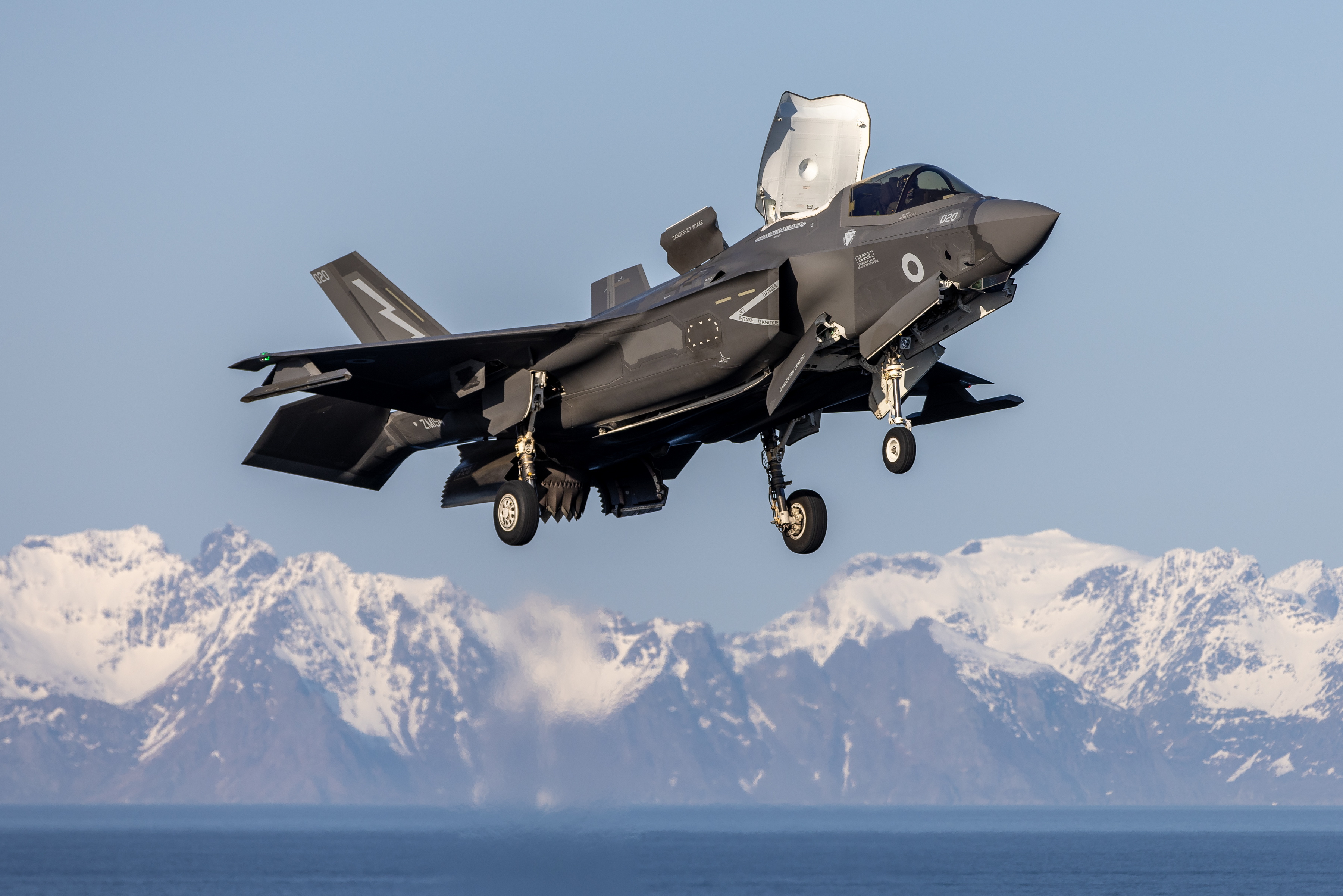 A UK F-35B hovering above an aircraft carrier, with snowy mountains in the background.