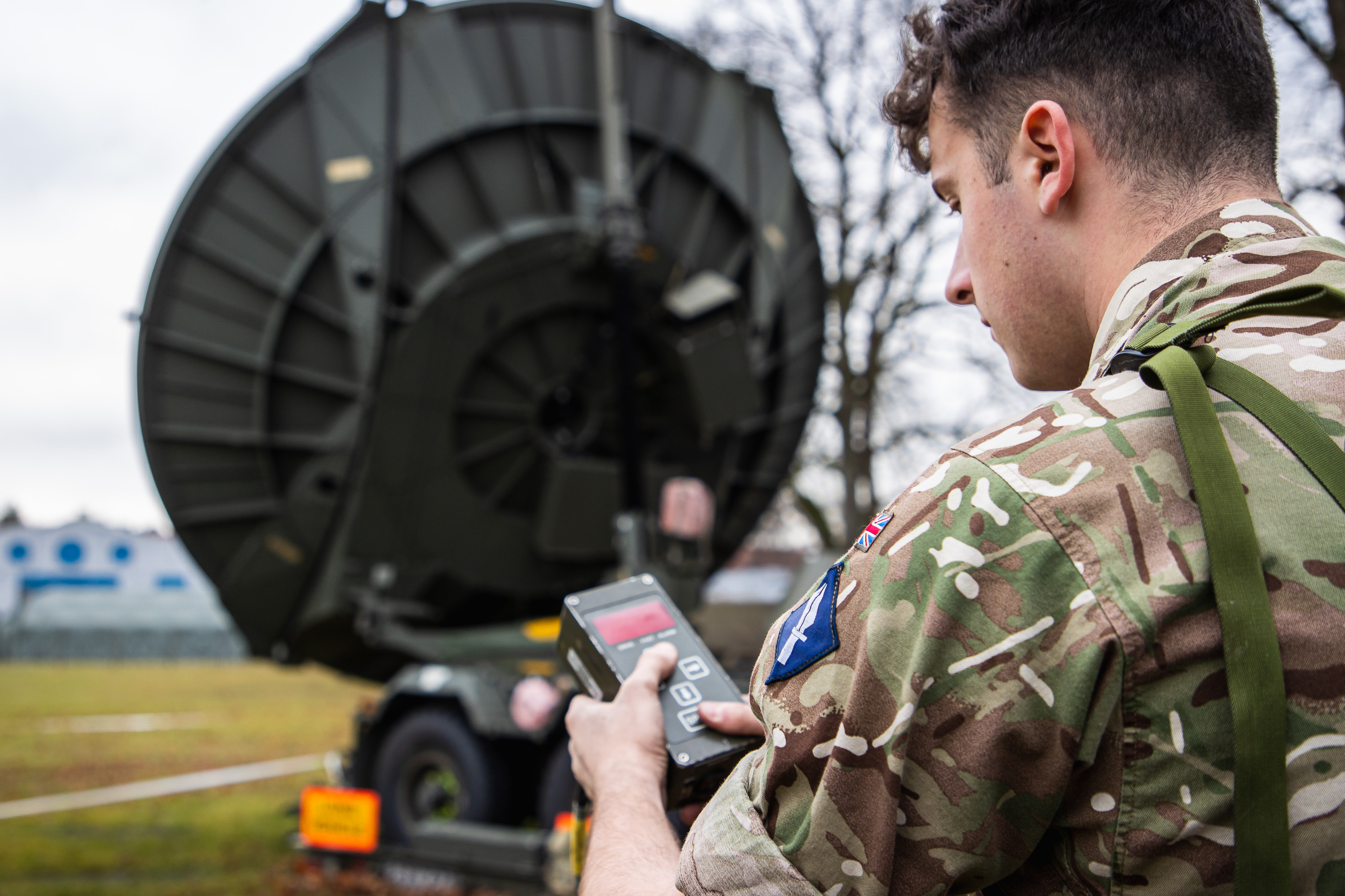 A British service person holding a remote in their hand. In the background is a satellite dish.