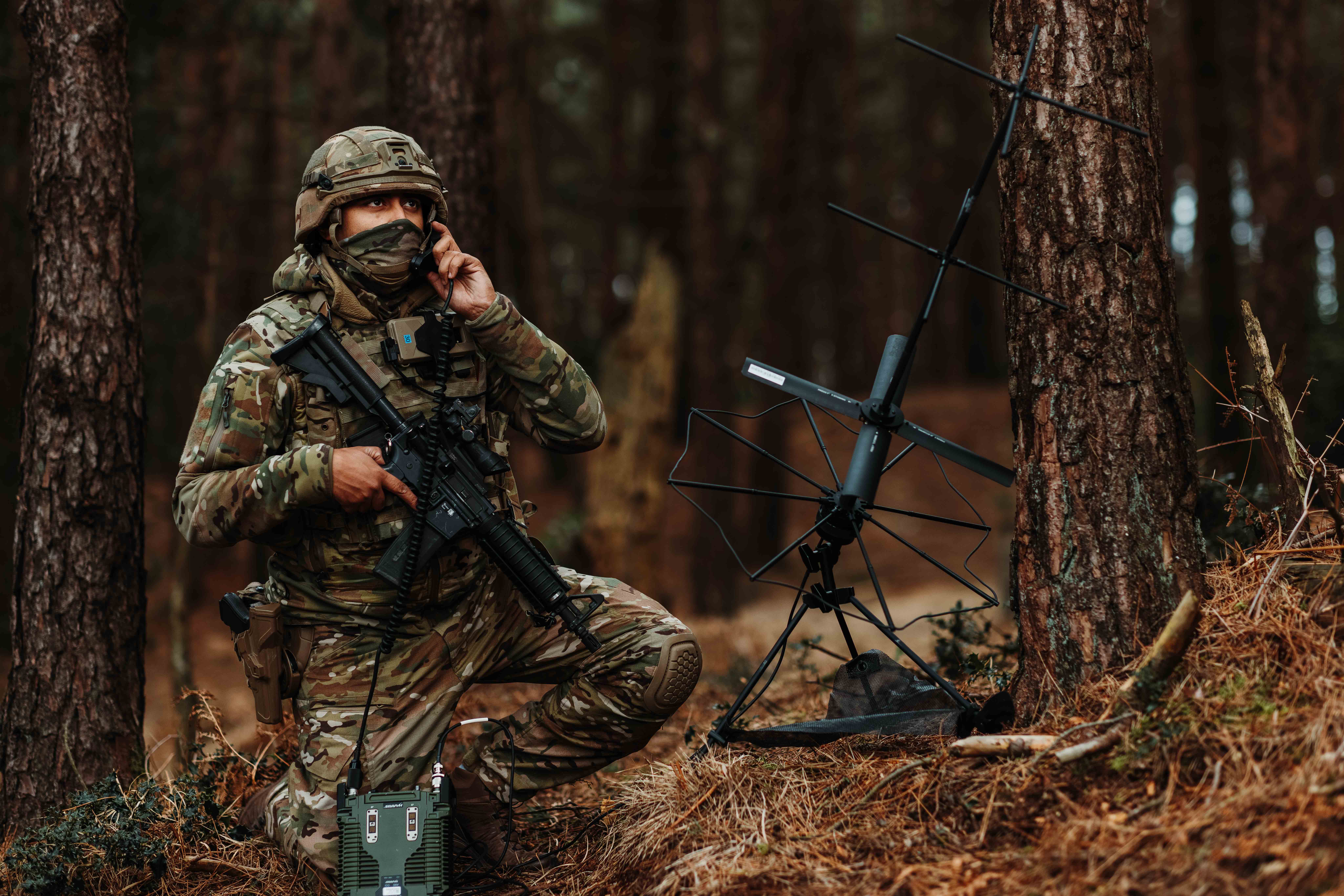 A military service person crouched in a forest using a field phone.