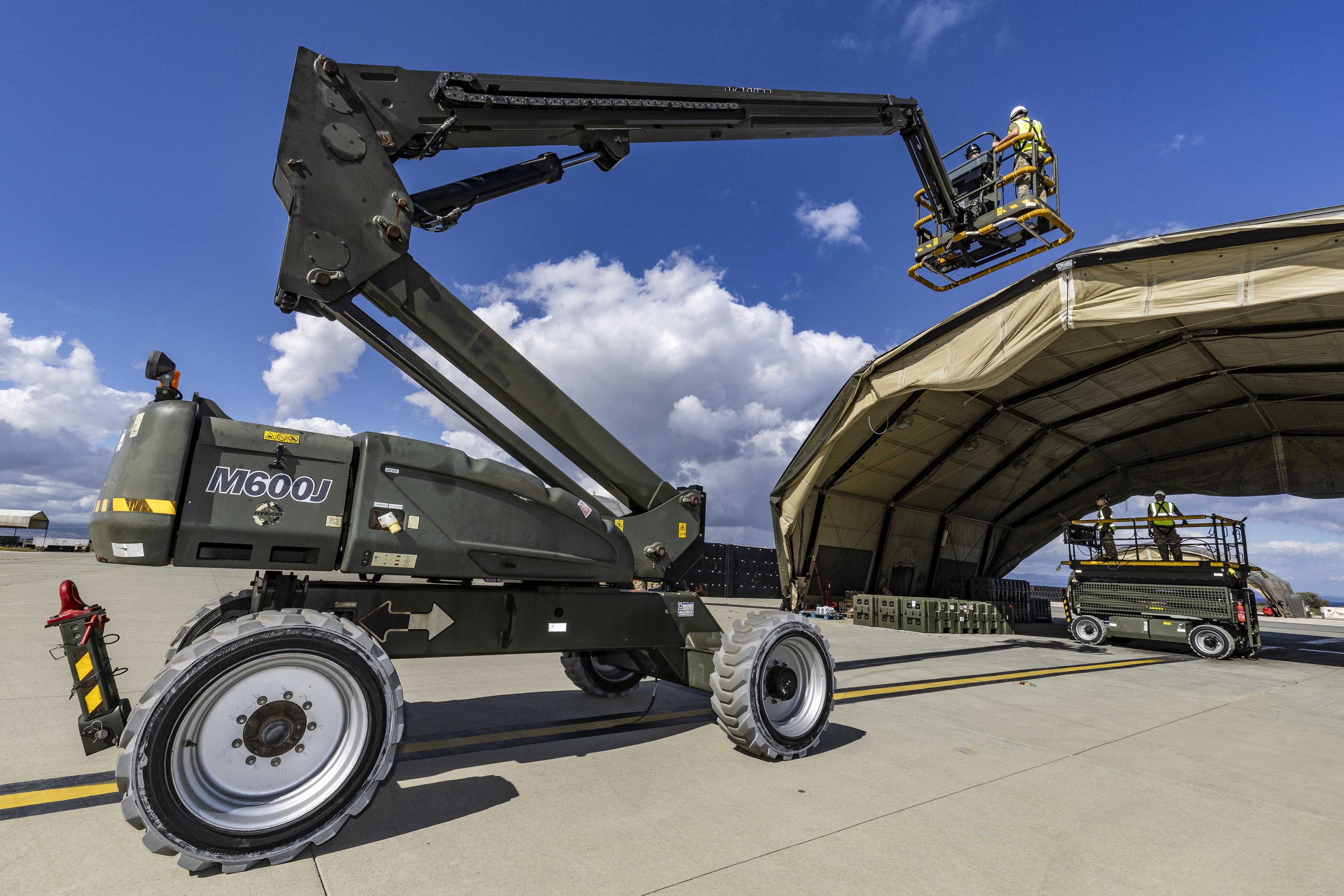 Close view of a boom lift raising personnel to the top edge of a temporary shelter, with additional workers and equipment positioned beneath the structure.