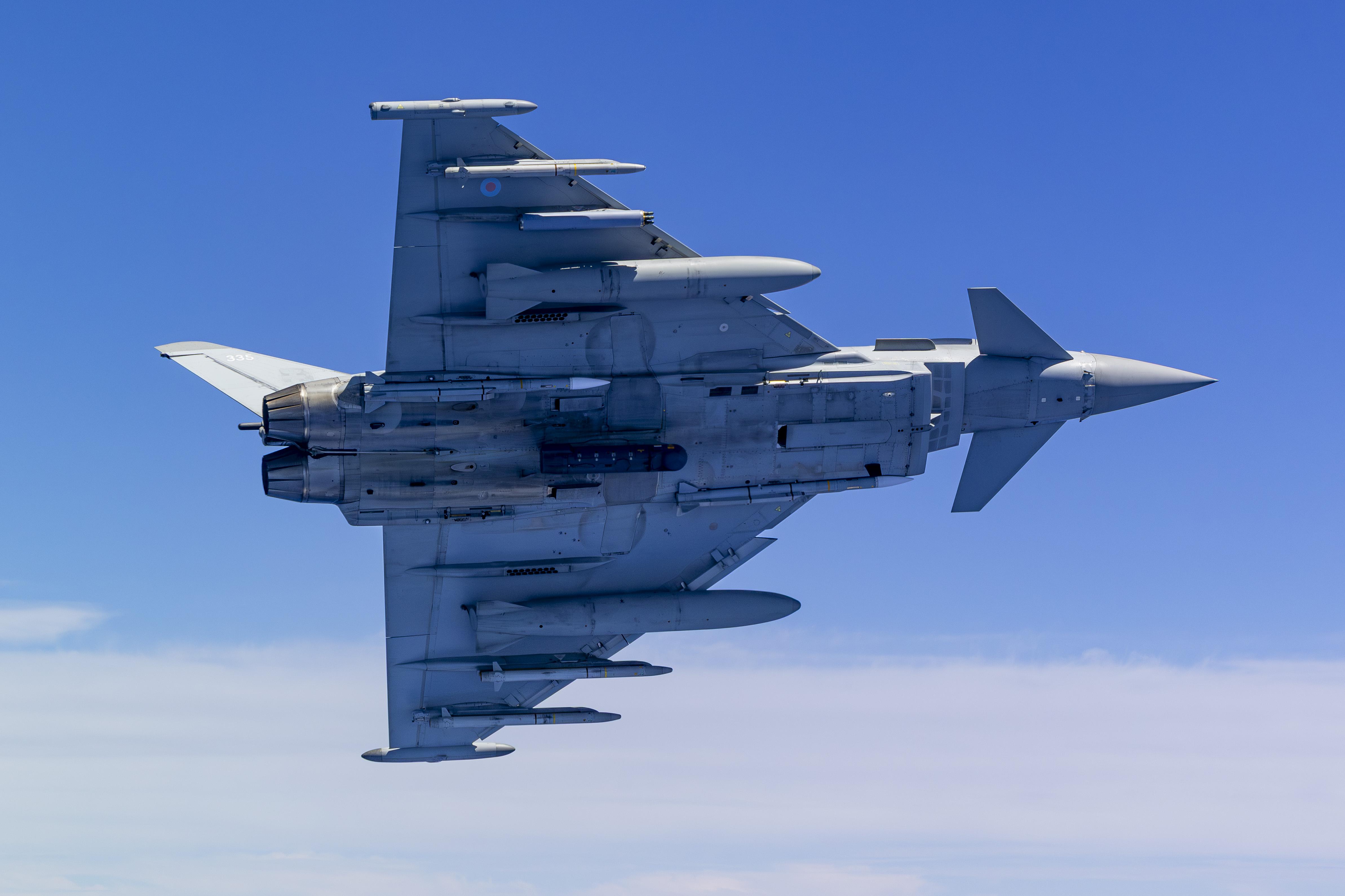 An RAF Typhoon banking vertically to the left as it flies through cloudy blue skies, allowing the APKWS fitted to its underside to clearly be seen.