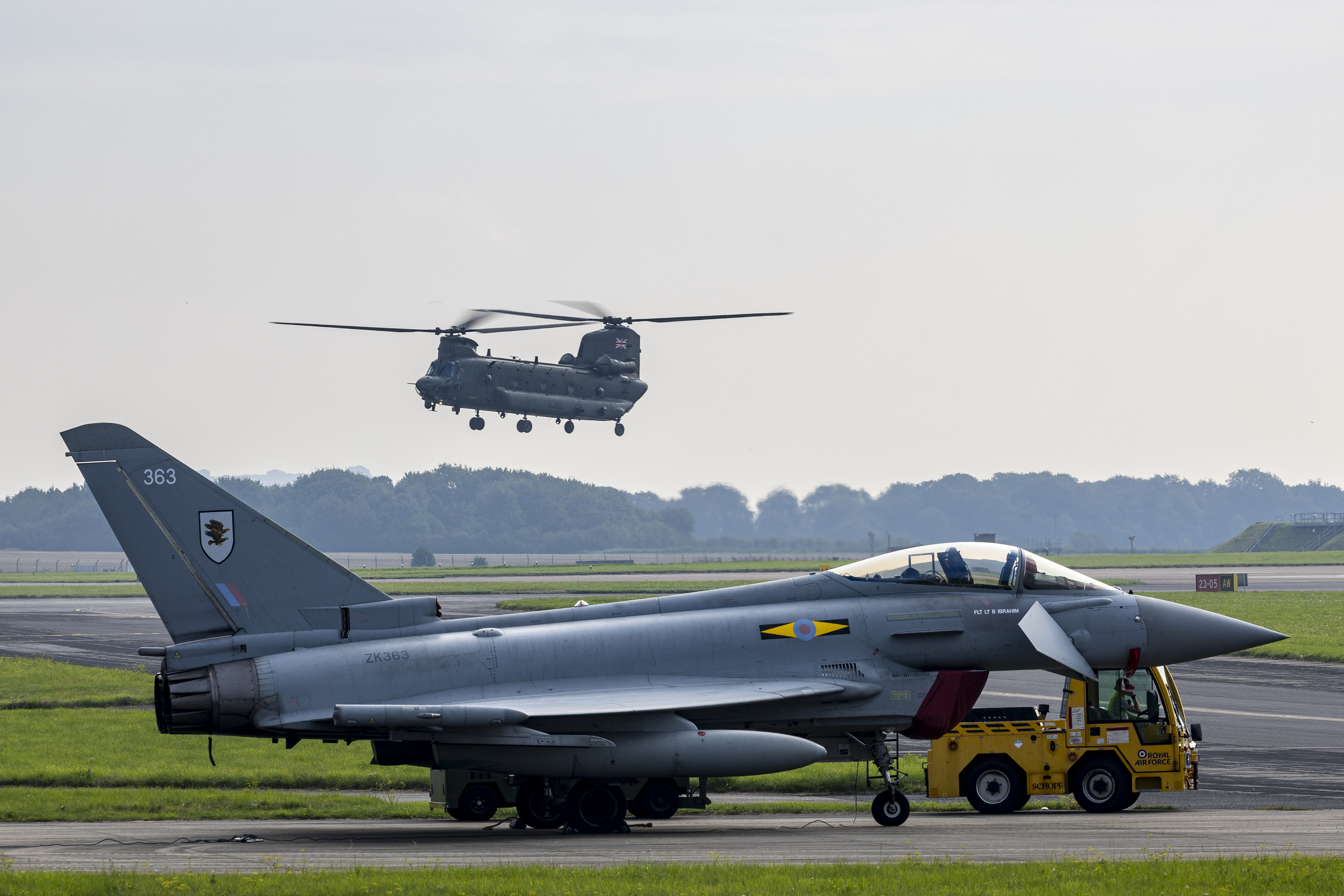 Image shows an RAF Typhoon aircraft on the ground with an RAF Chinook flying low level in the background.
