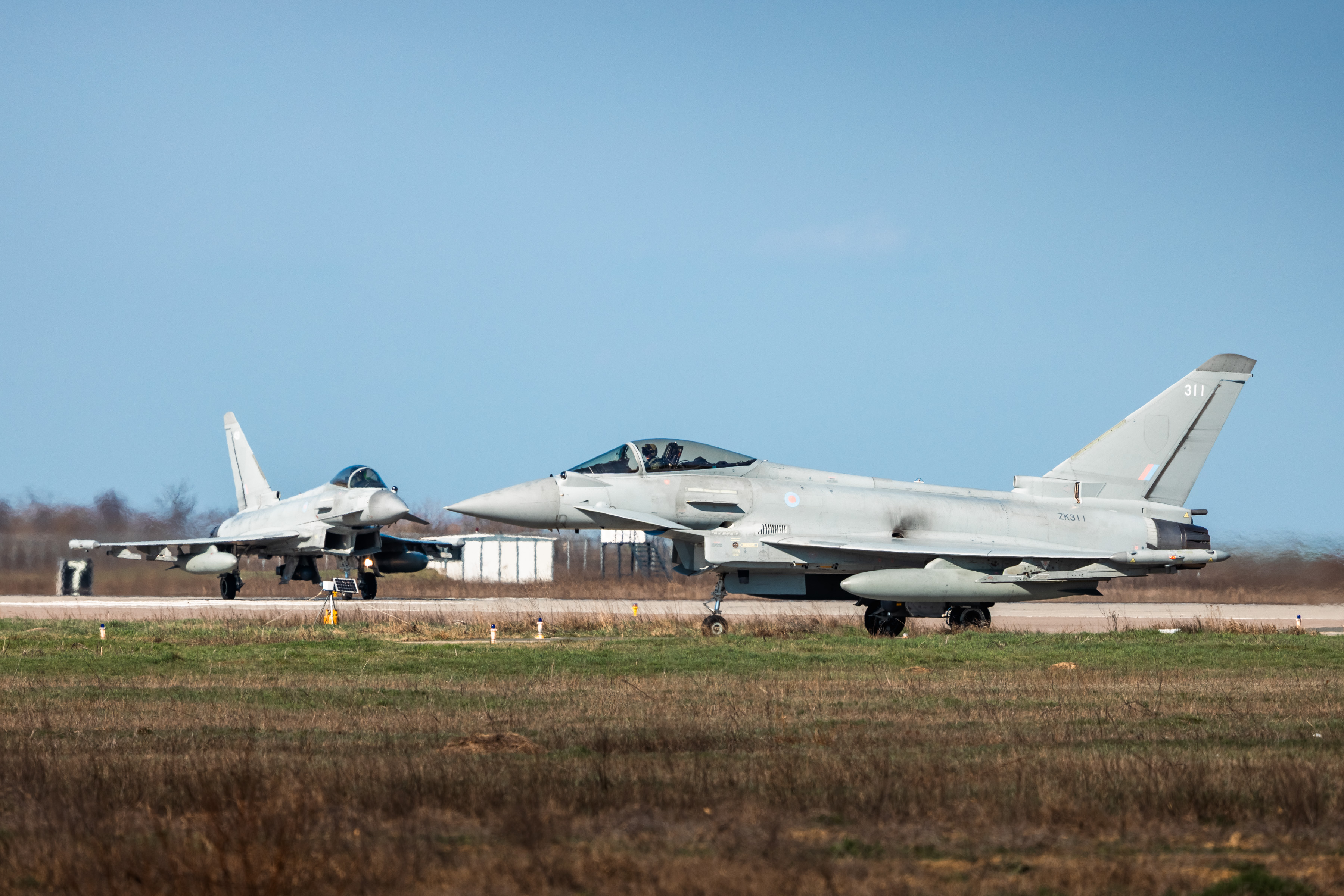 Two RAF Typhoons on a runway, one taxiing in the foreground and another approaching or landing in the background.