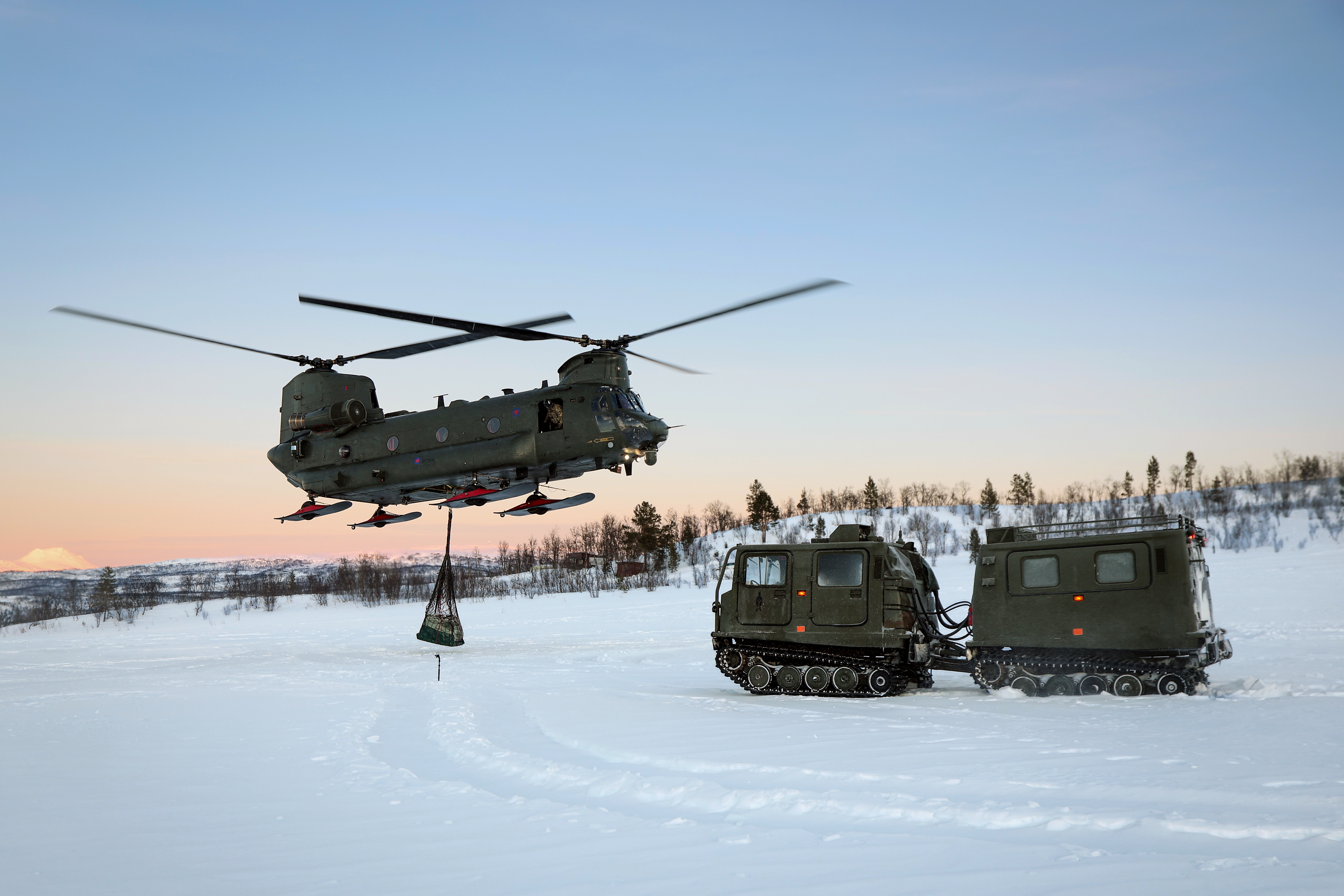 A Chinook hovering over snowy terrain in the High North with skis fitted. On the ground are two armoured vehicles on treads.