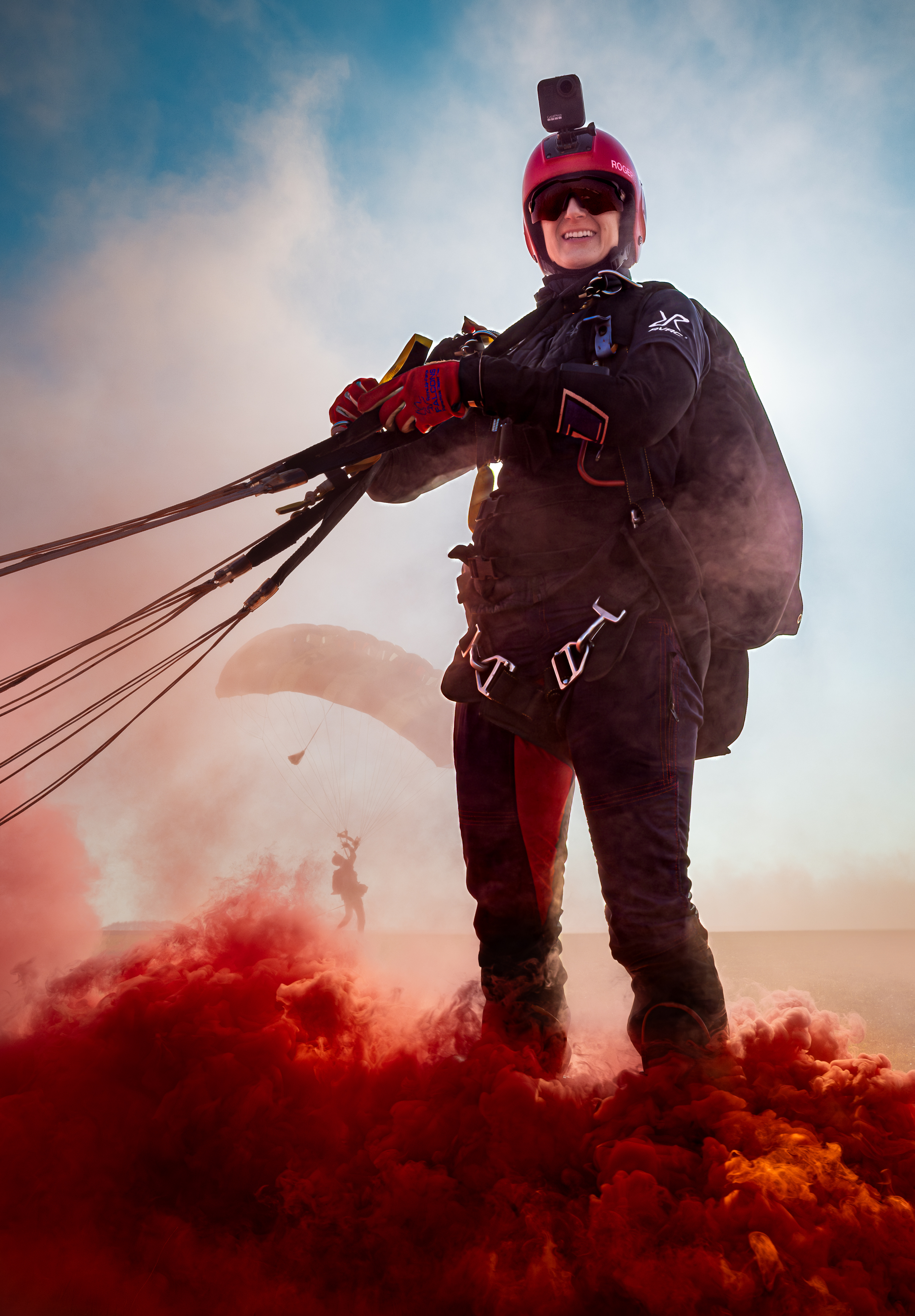 A parachutist on the ground still attached to a parachute with a red smoke flair billowing around them