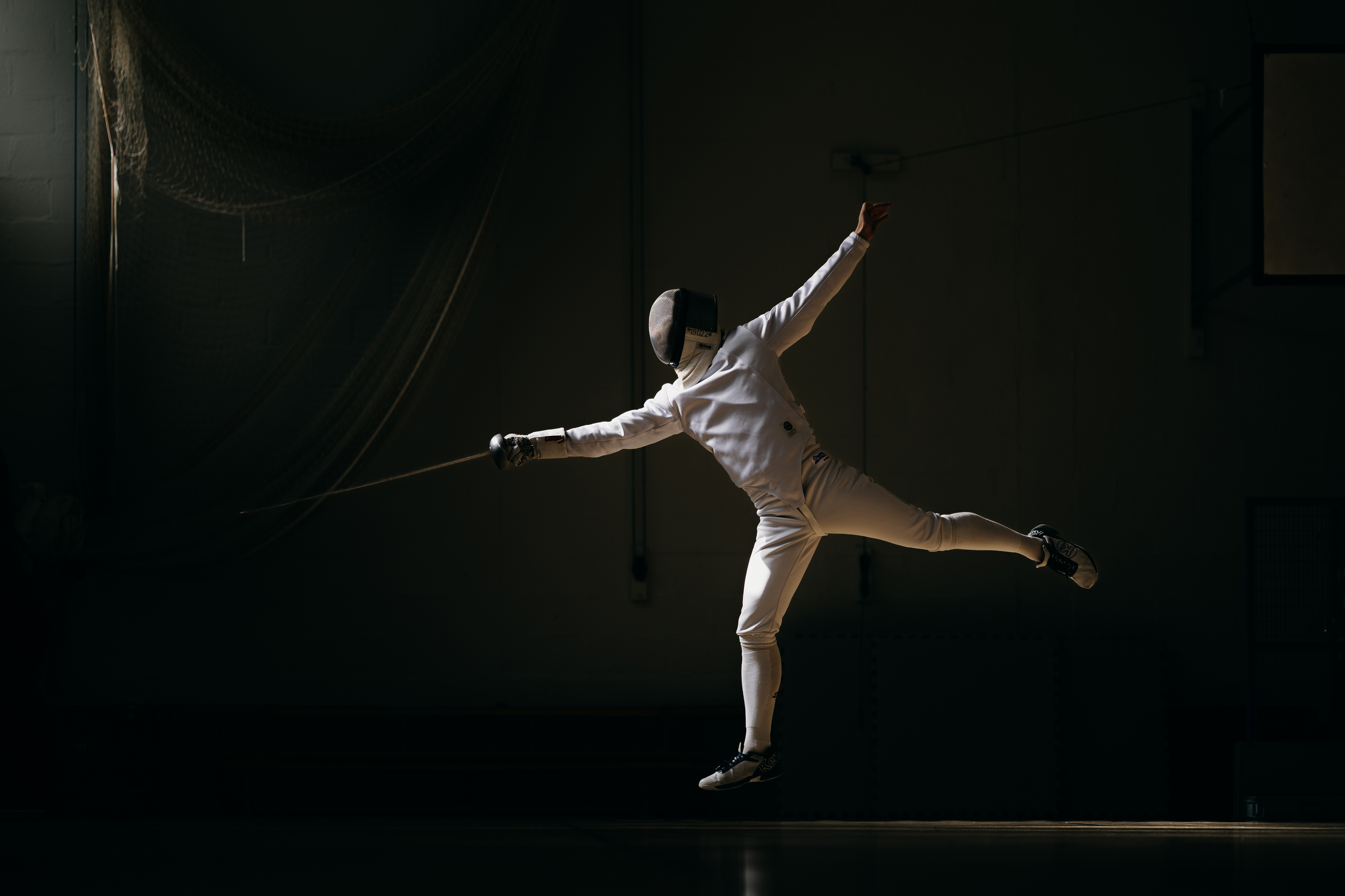A fencer contrasting against a black background in their white fencing clothing with foil out stretched