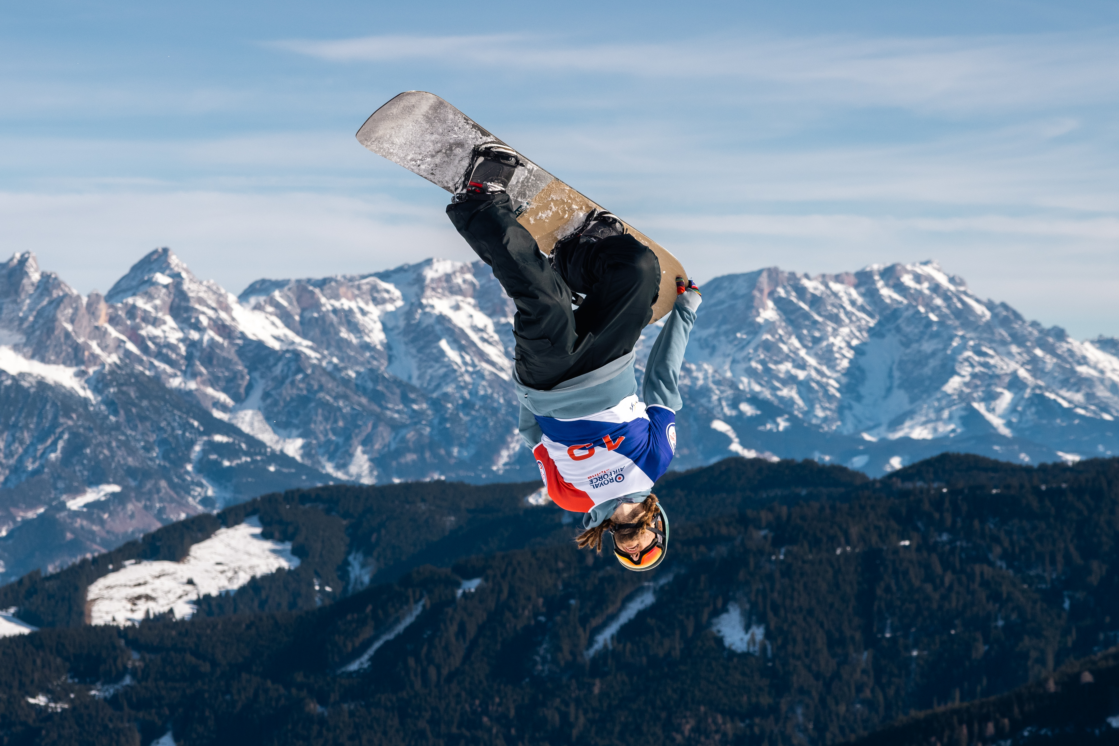 A snow boarder upside down in the air with snow topped mountains in the background