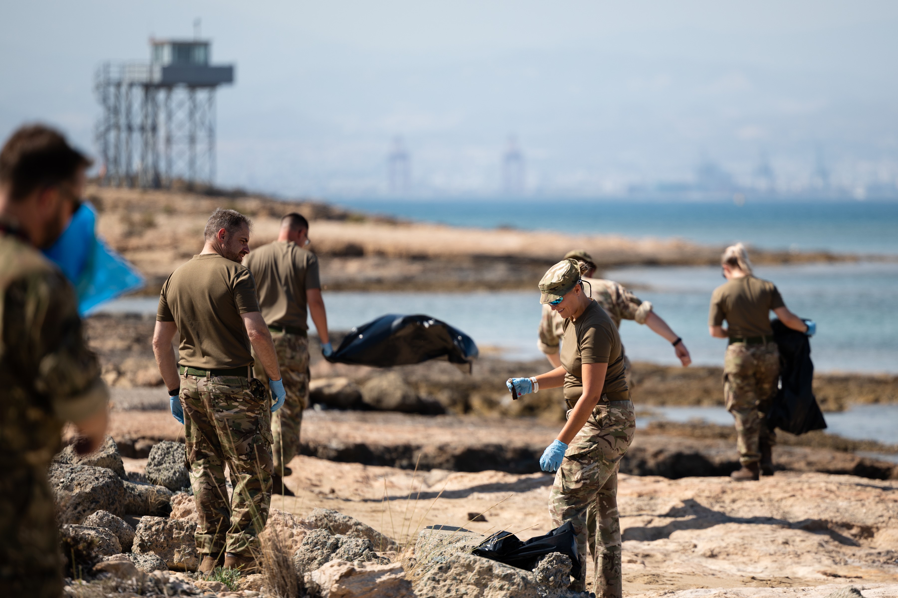 Six servicepeople cleaning up on the beach, with bin bags and gloves, in a rocky area.