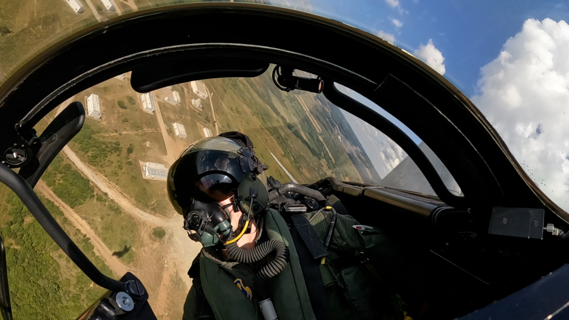 A pilot in the cockpit of a fast jet, banking over grassy land beneath.