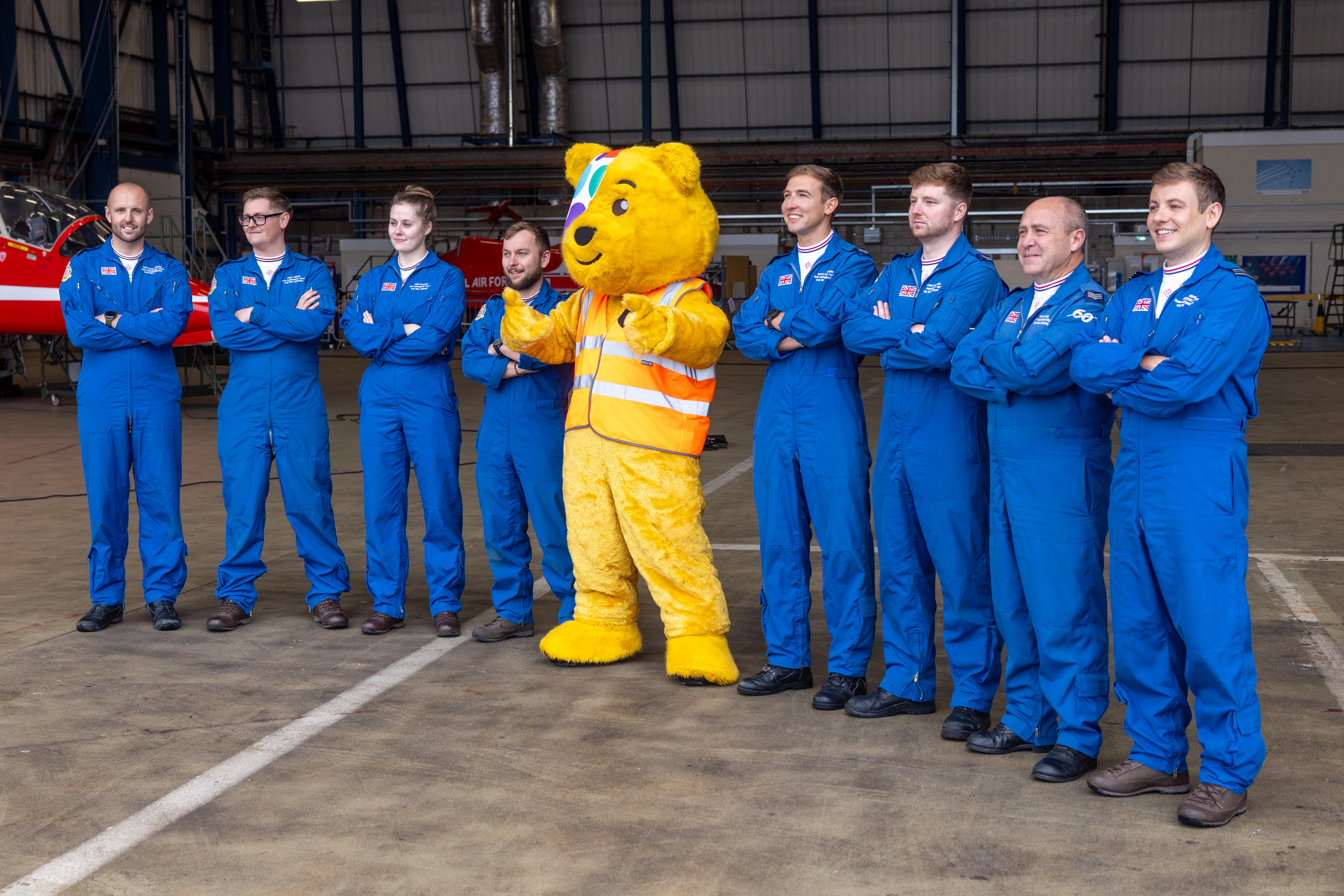 Pudsey the Children in Need mascot stands next to members of the Red Arrows' support team - dressed in their distinctive blue coveralls - inside the hangar at RAF Waddington.